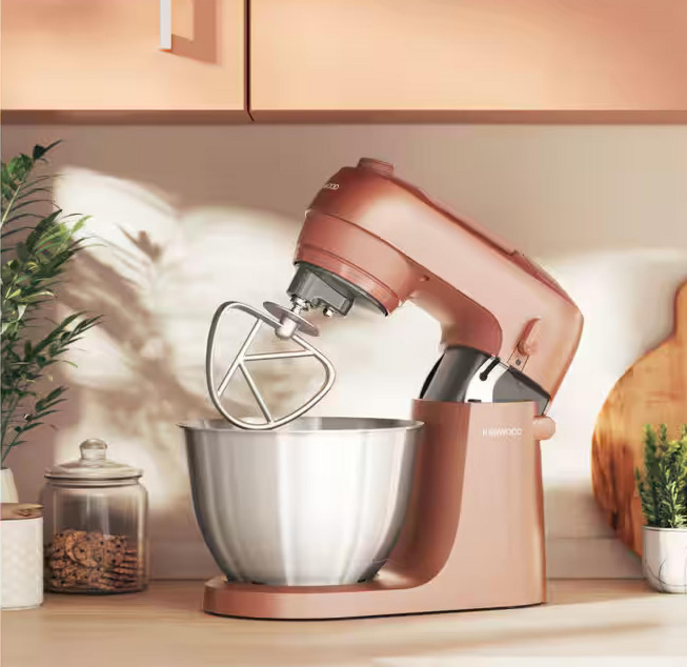 Ein rosafarbener Kenwood-Küchenmixer mit einer Edelstahlschüssel auf einer Holzoberfläche. Pflanzen und Gläser sind im Hintergrund.