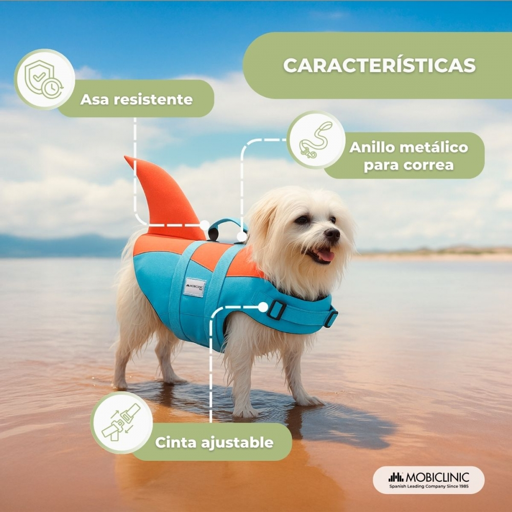 Un perro blanco pequeño lleva un chaleco salvavidas con aleta de tiburón azul y naranja de pie en agua poco profunda en la playa.