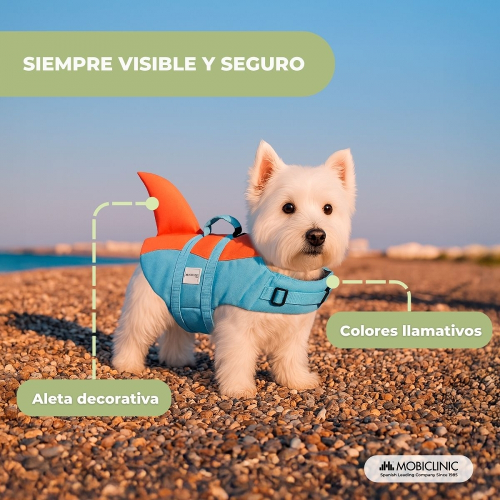 Un perro blanco lleva un chaleco salvavidas azul y naranja con aleta de tiburón. Está en una playa de guijarros con cielo azul.