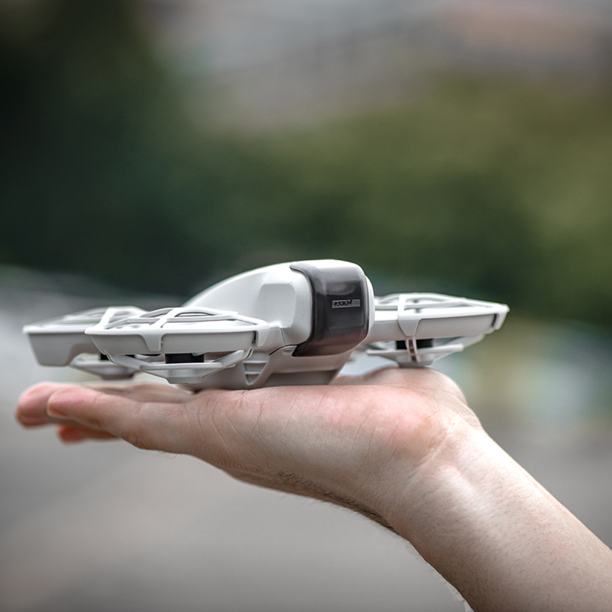 Biały dron trzymany w dłoni osoby. Dron ma cztery śmigła. Tło jest rozmyte z zielonymi i szarymi kolorami.