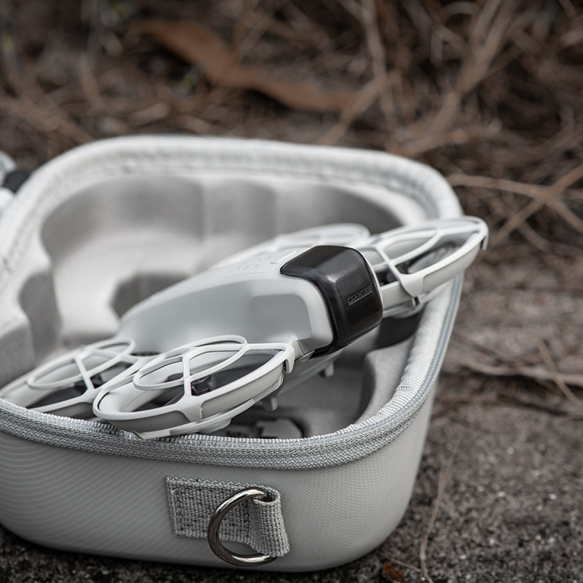 Szary dron spoczywa w jasnoszarej walizce. Walizka stoi na piaszczystej powierzchni z suchymi gałązkami w tle.