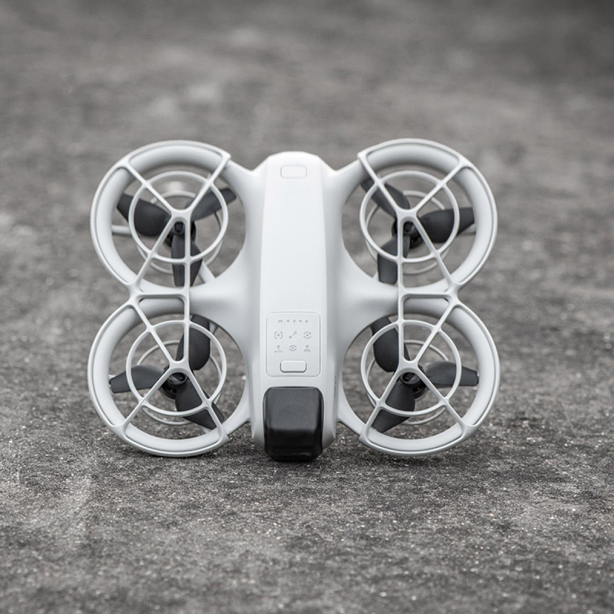 Biały dron z czterema śmigłami, czarnymi łopatkami i czarną kamerą na szarej betonowej powierzchni.