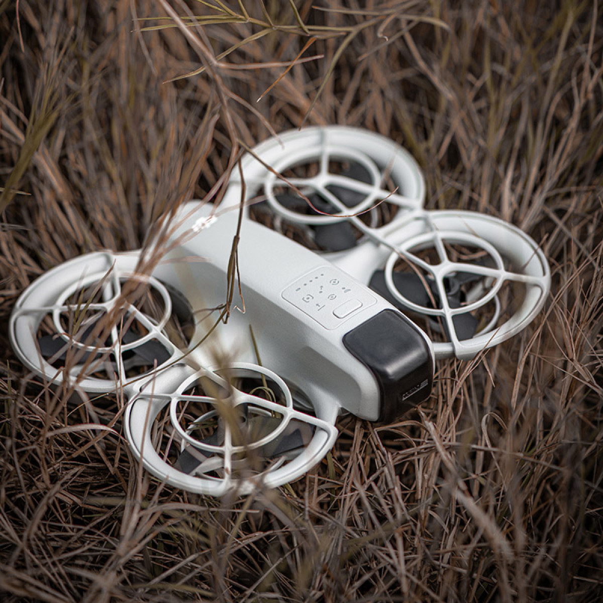 Biały dron z czarnymi akcentami spoczywa w suchej trawie. Dron ma cztery śmigła i jest skierowany w stronę widza.