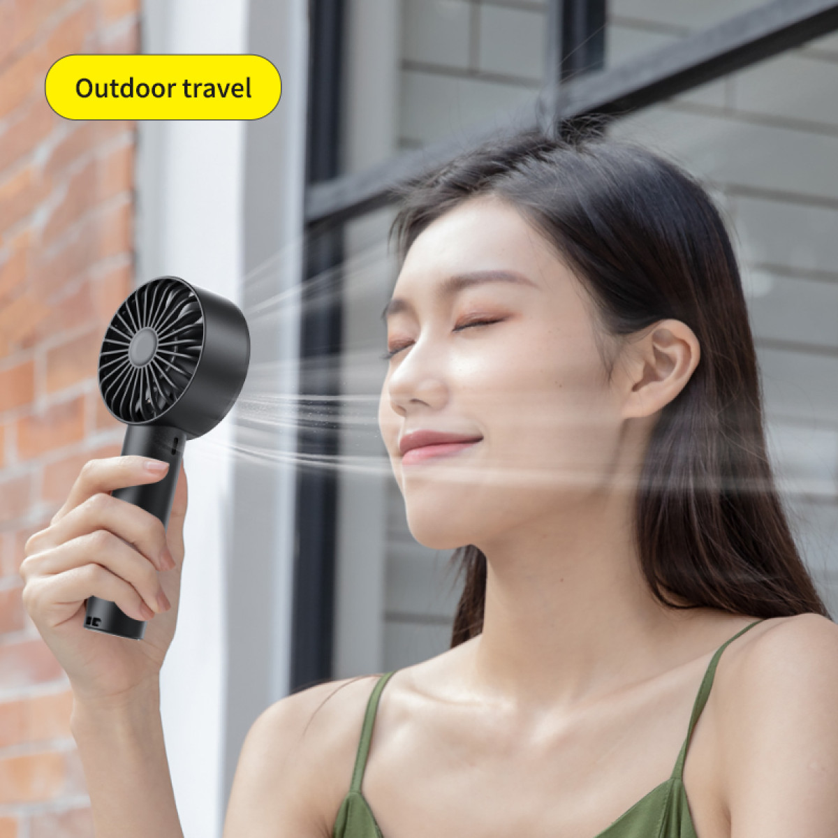 Kobieta cieszy się powiewem z czarnego wentylatora ręcznego. Wentylator jest blisko jej twarzy. Ma długie, ciemne włosy i zielony top.
