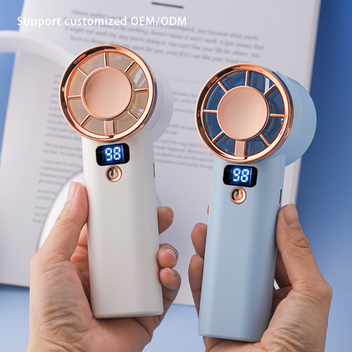 Dwa wentylatory ręczne, jeden biały, jeden niebieski, trzymane w dłoniach. Oba mają wyświetlacze cyfrowe z '98'. Tło to papier.