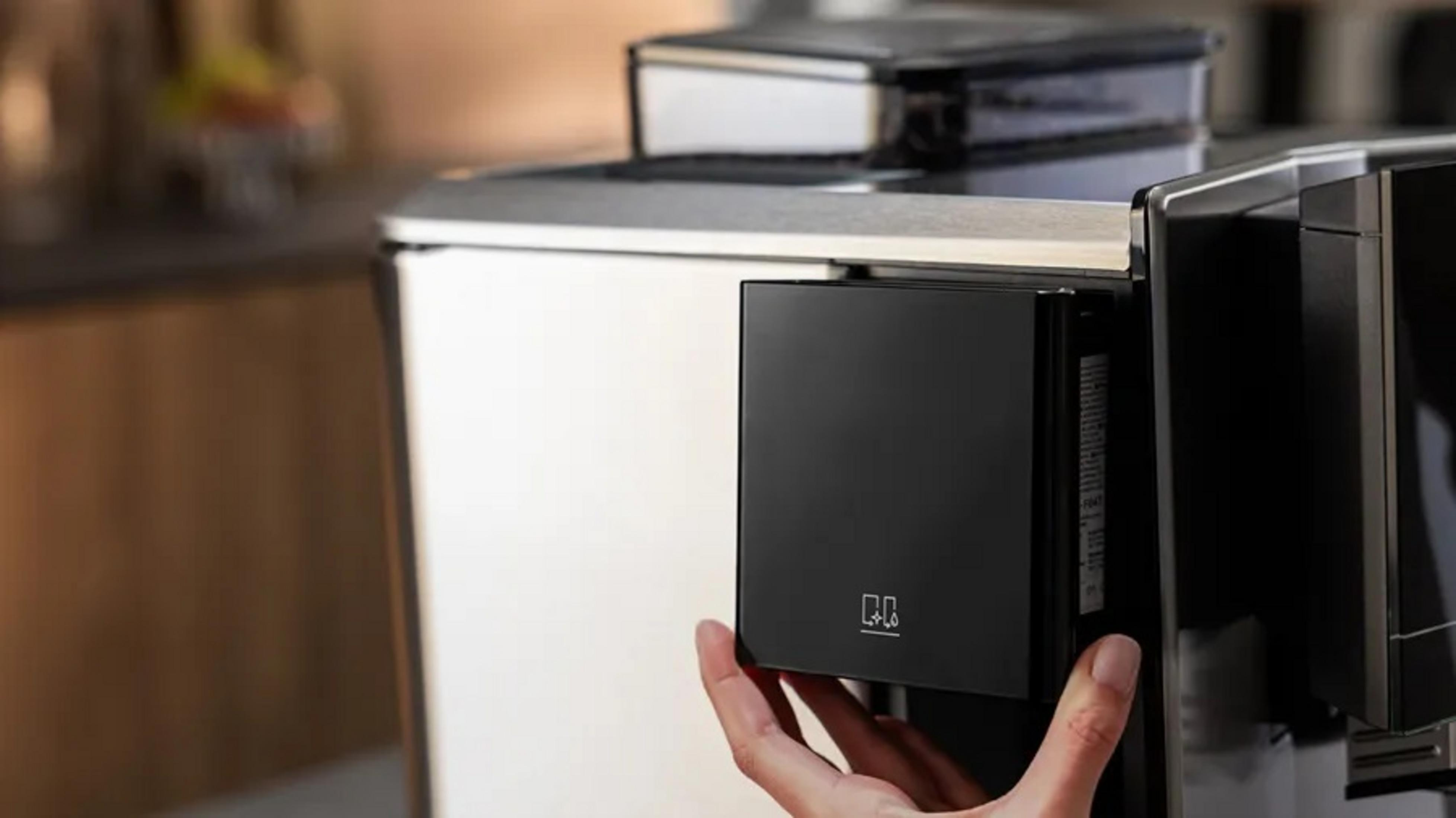 Ręka trzyma czarny element ekspresu do kawy. Maszyna jest srebrna i czarna. Tło to rozmyta kuchnia.