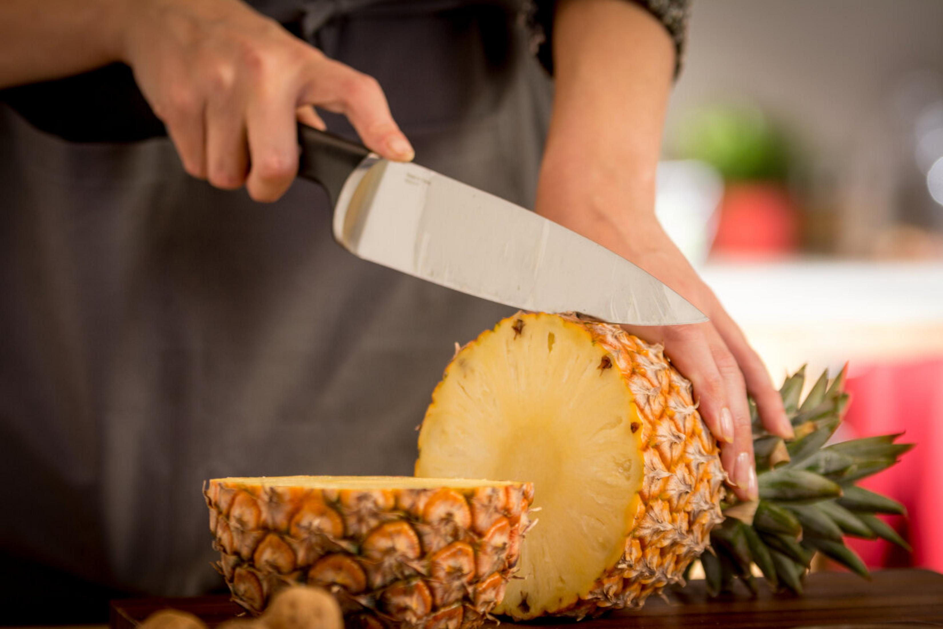 Una persona corta una piña por la mitad con un cuchillo grande. La fruta está sobre madera.