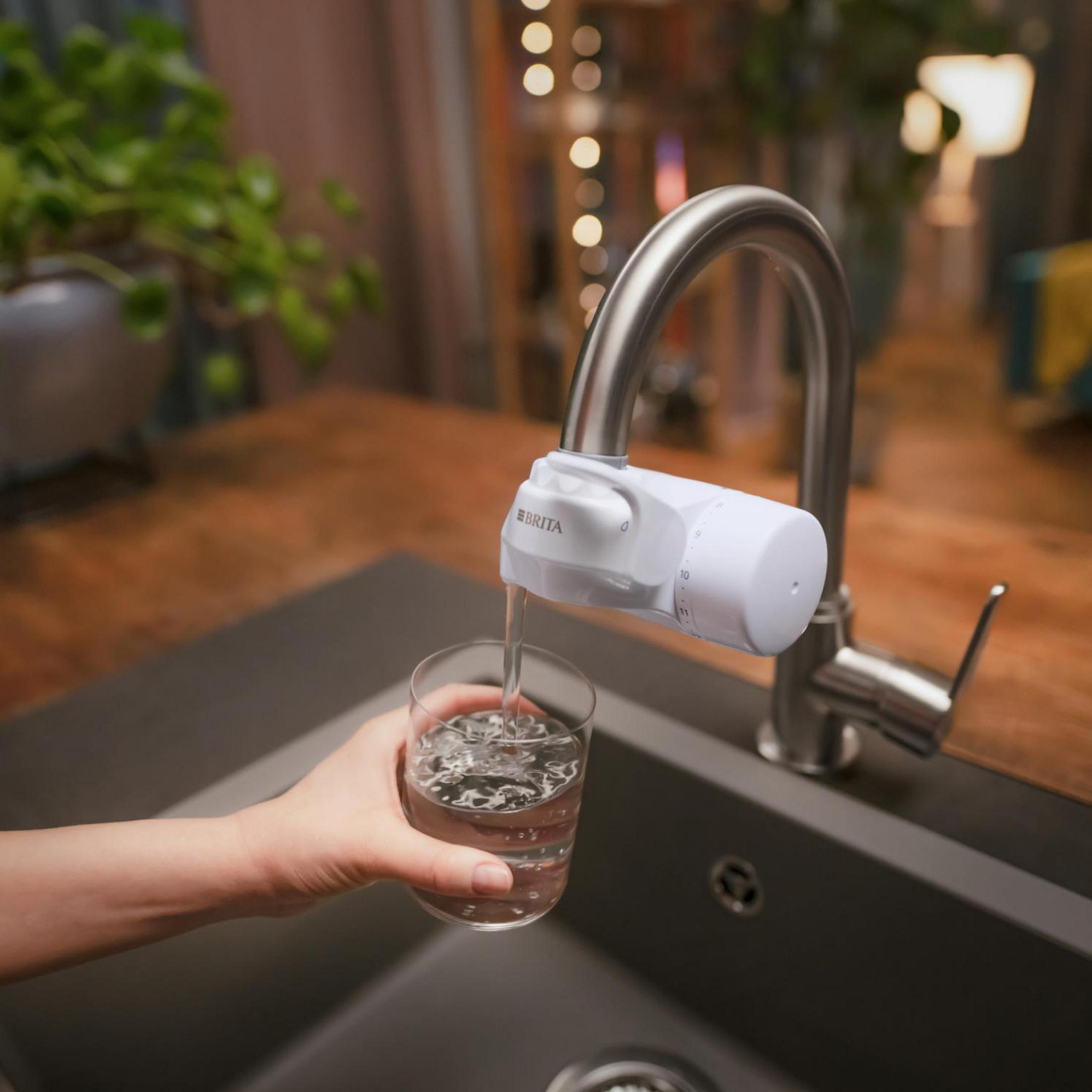 Una mano tiene un bicchiere mentre l'acqua scorre da un rubinetto con filtro Brita, sopra un lavandino scuro.
