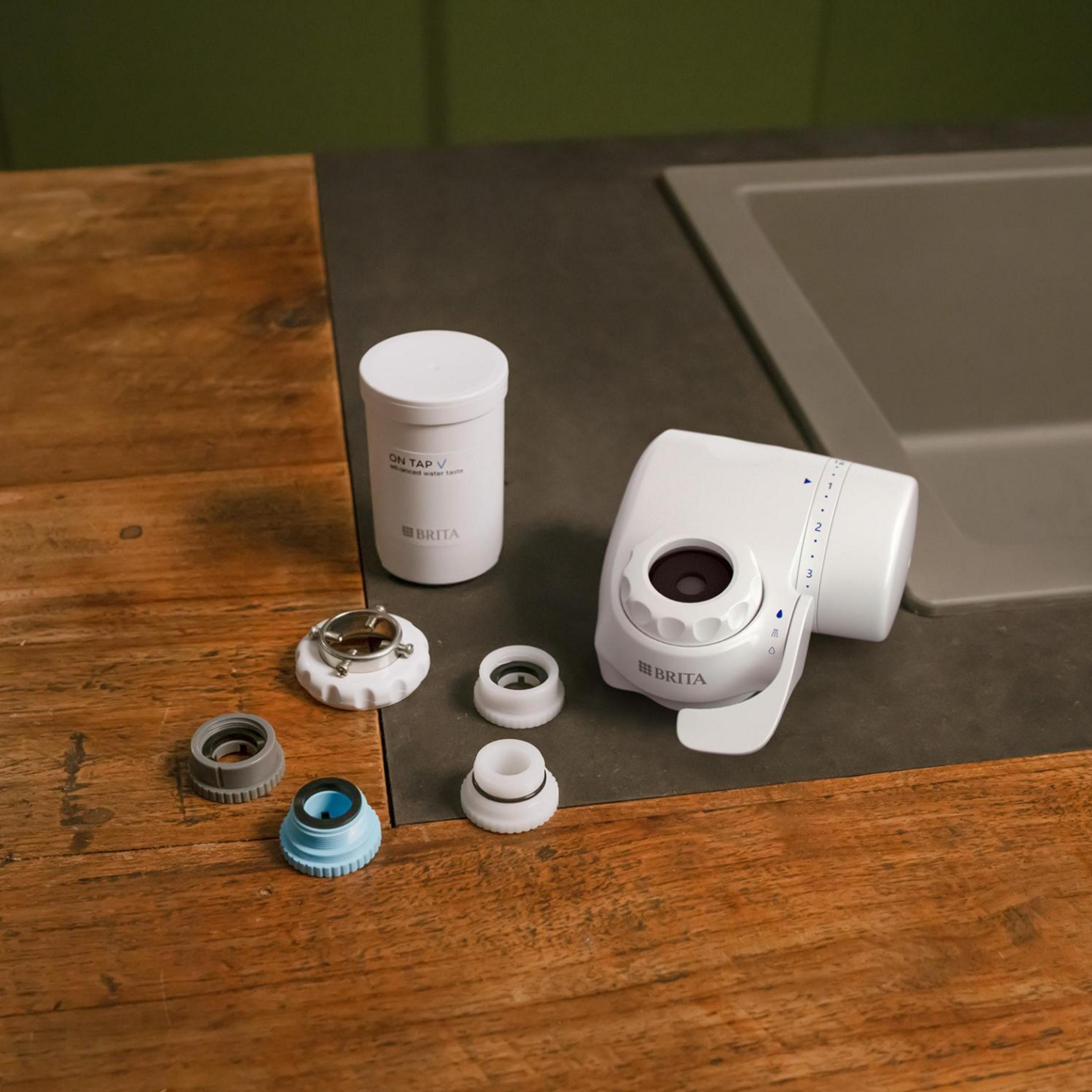 Un sistema di filtraggio dell'acqua Brita bianco con varie parti su una superficie di legno vicino a un lavandino e sfondo verde.