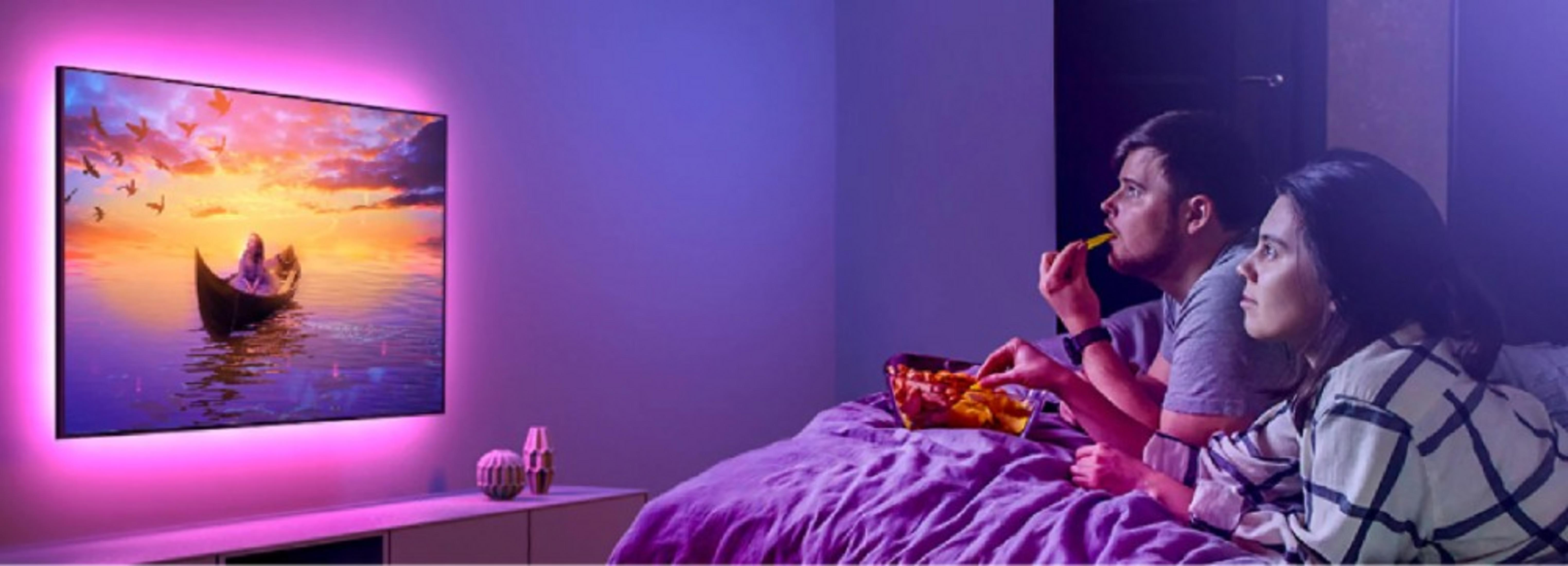 Coppia guarda la TV. L'uomo mangia uno snack. La TV mostra una scena di barca. Luce ambientale viola e rosa.