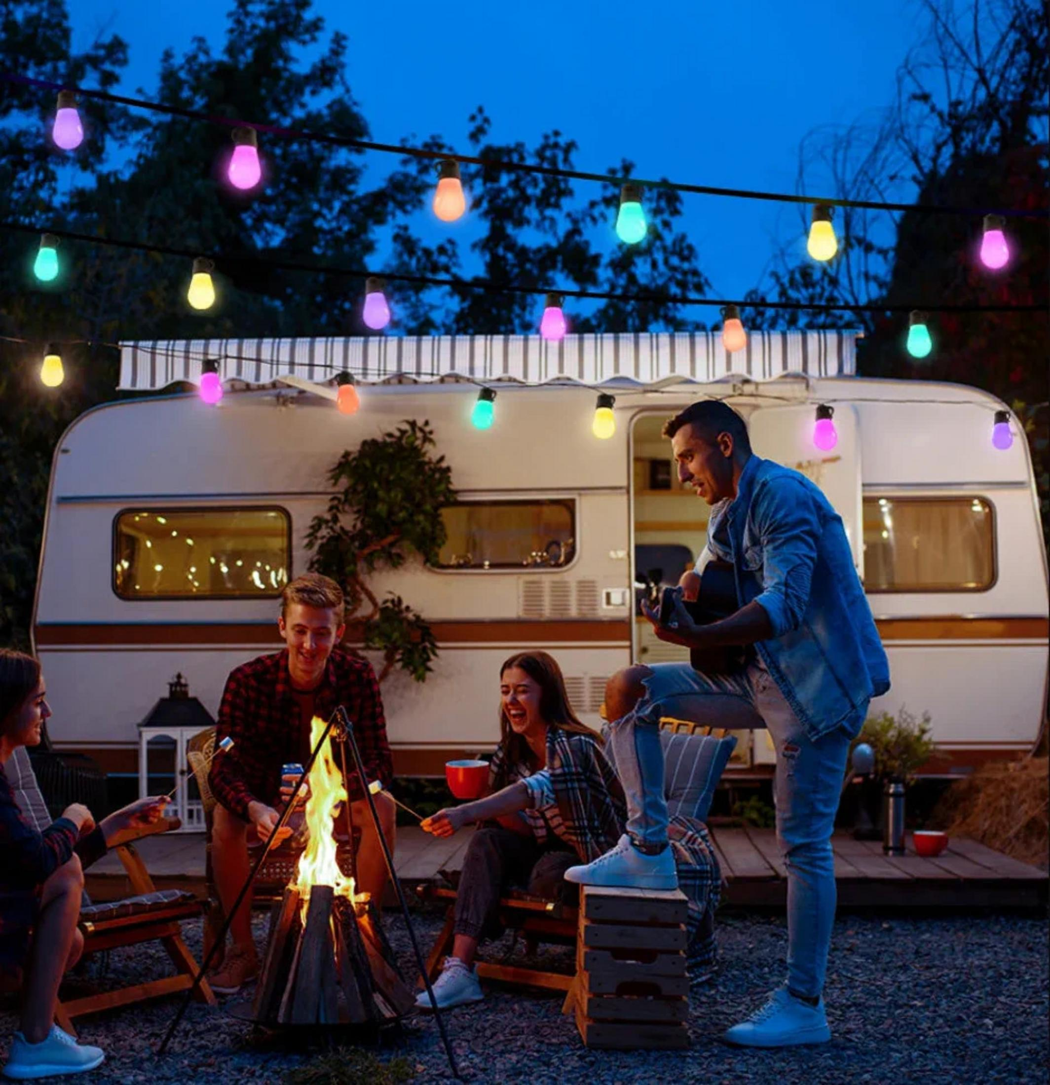 Przyjaciele wokół ogniska z przyczepą kempingową w tle, światła nad, jeden gra na gitarze.