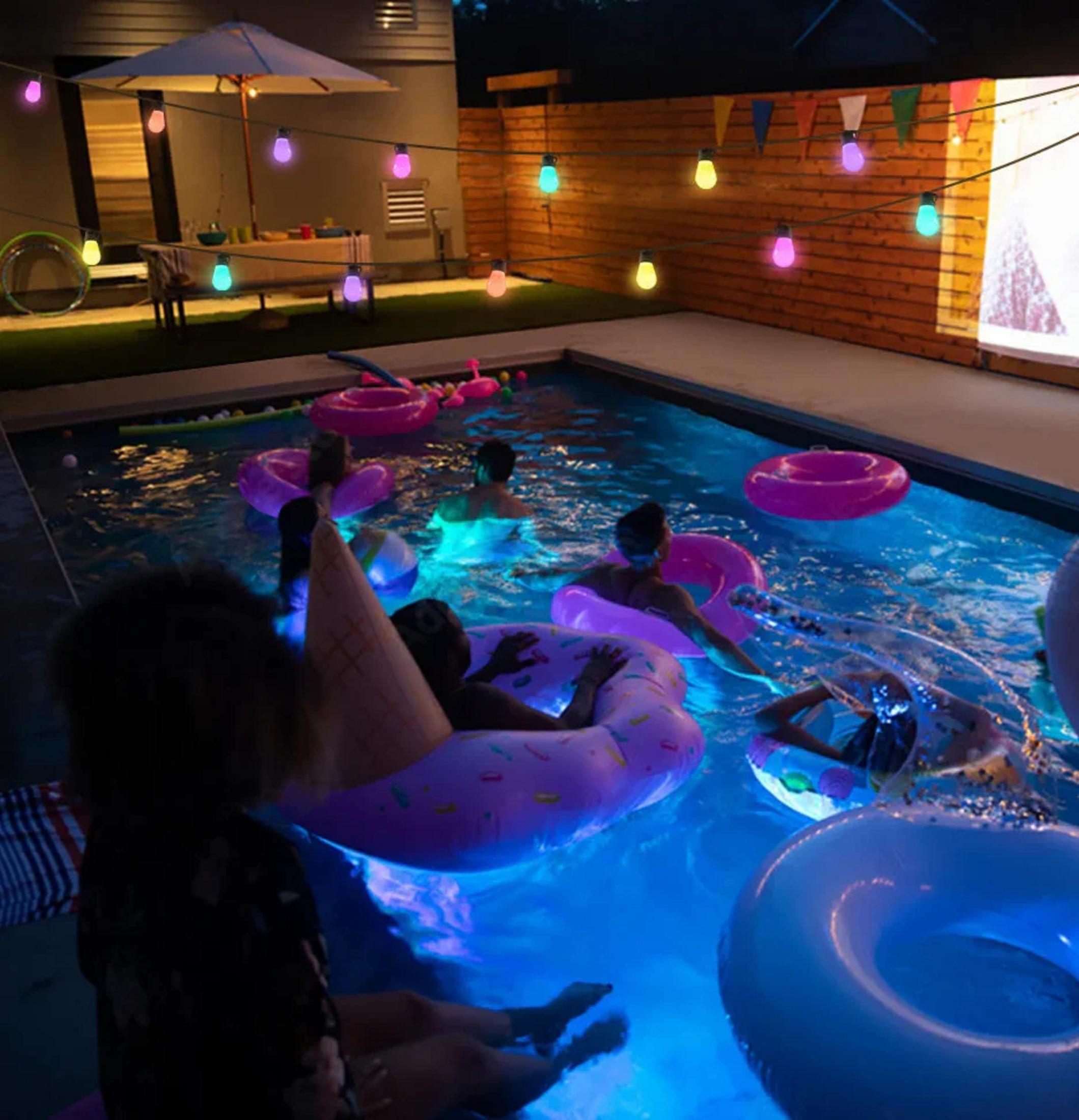 Ludzie w basenie w nocy z kolorowymi światłami, kołami do pływania. Niektórzy są w basenie, inni siedzą.