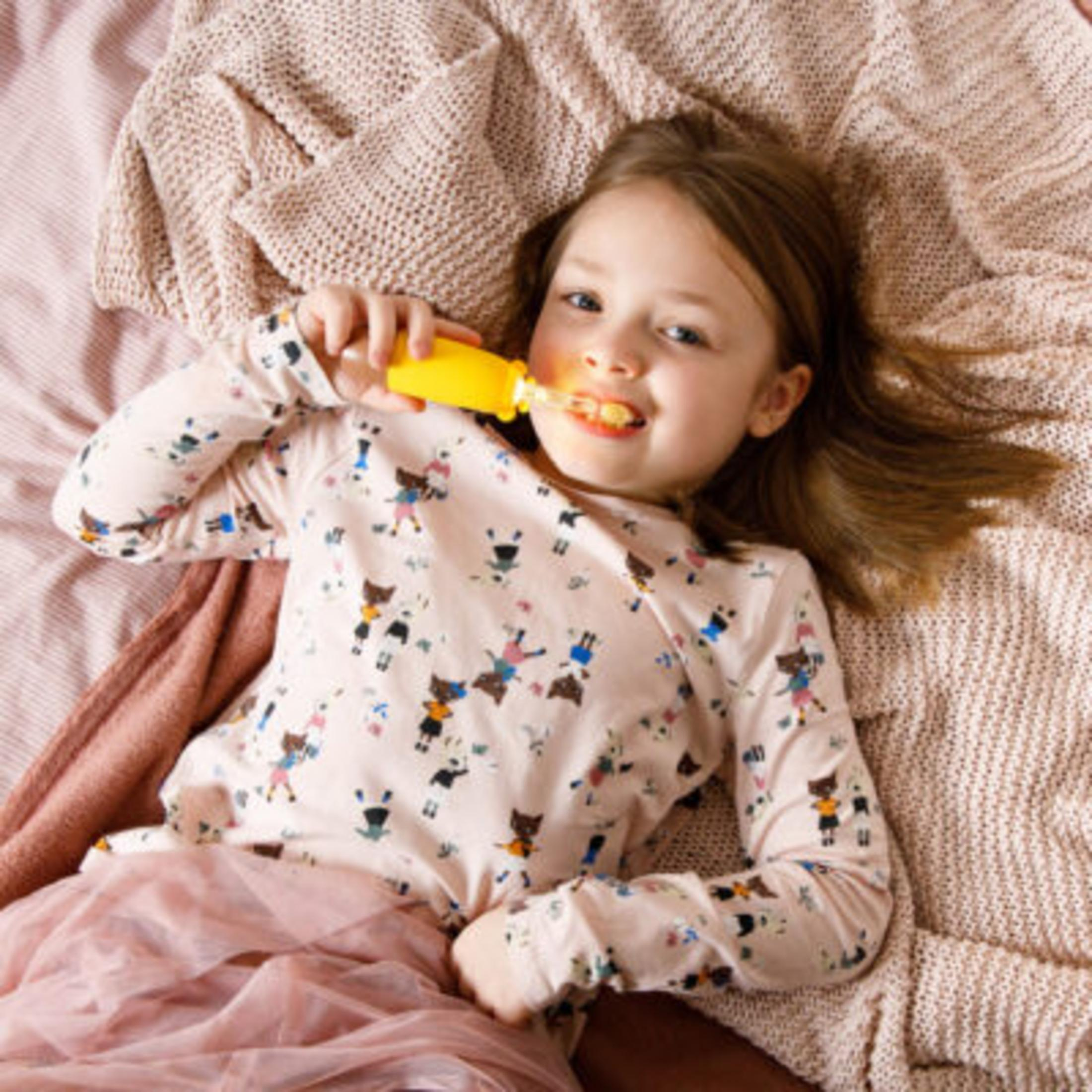 Una niña está acostada en una cama, cepillándose los dientes con un cepillo de dientes amarillo. Lleva una camisa estampada y una falda rosa.