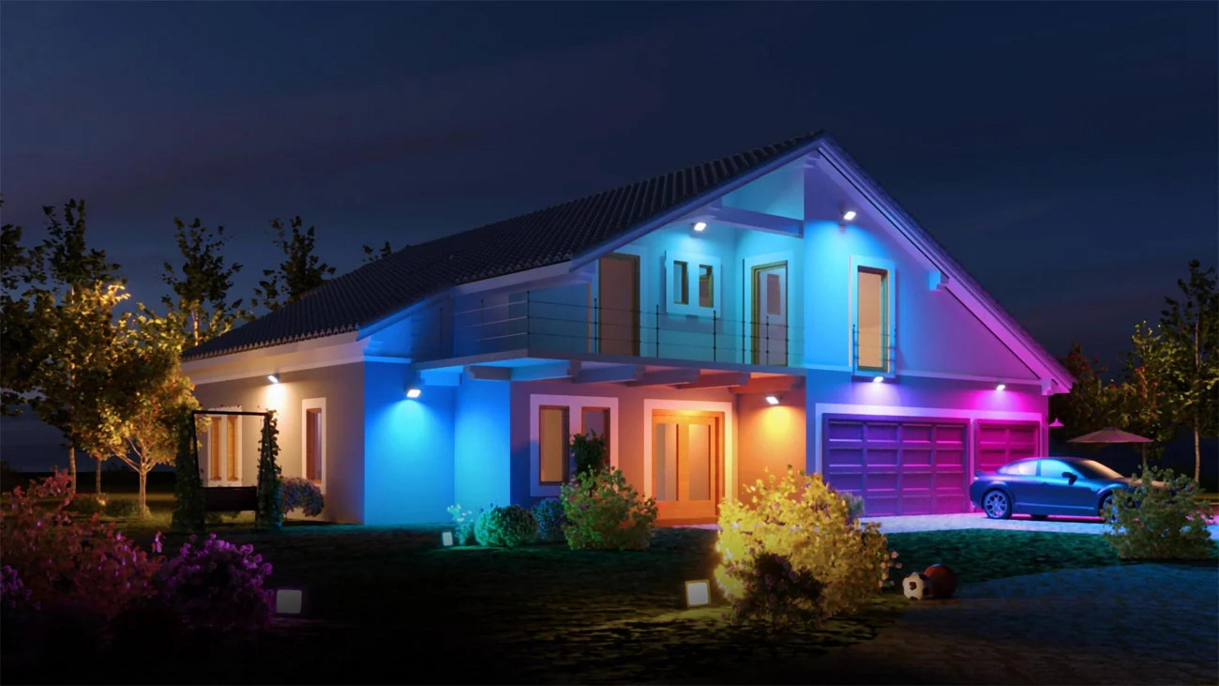 Nowoczesny dom oświetlony kolorowymi światłami. Samochód zaparkowany na podjeździe. Drzewa i krzewy otaczają dom pod ciemnym niebem.