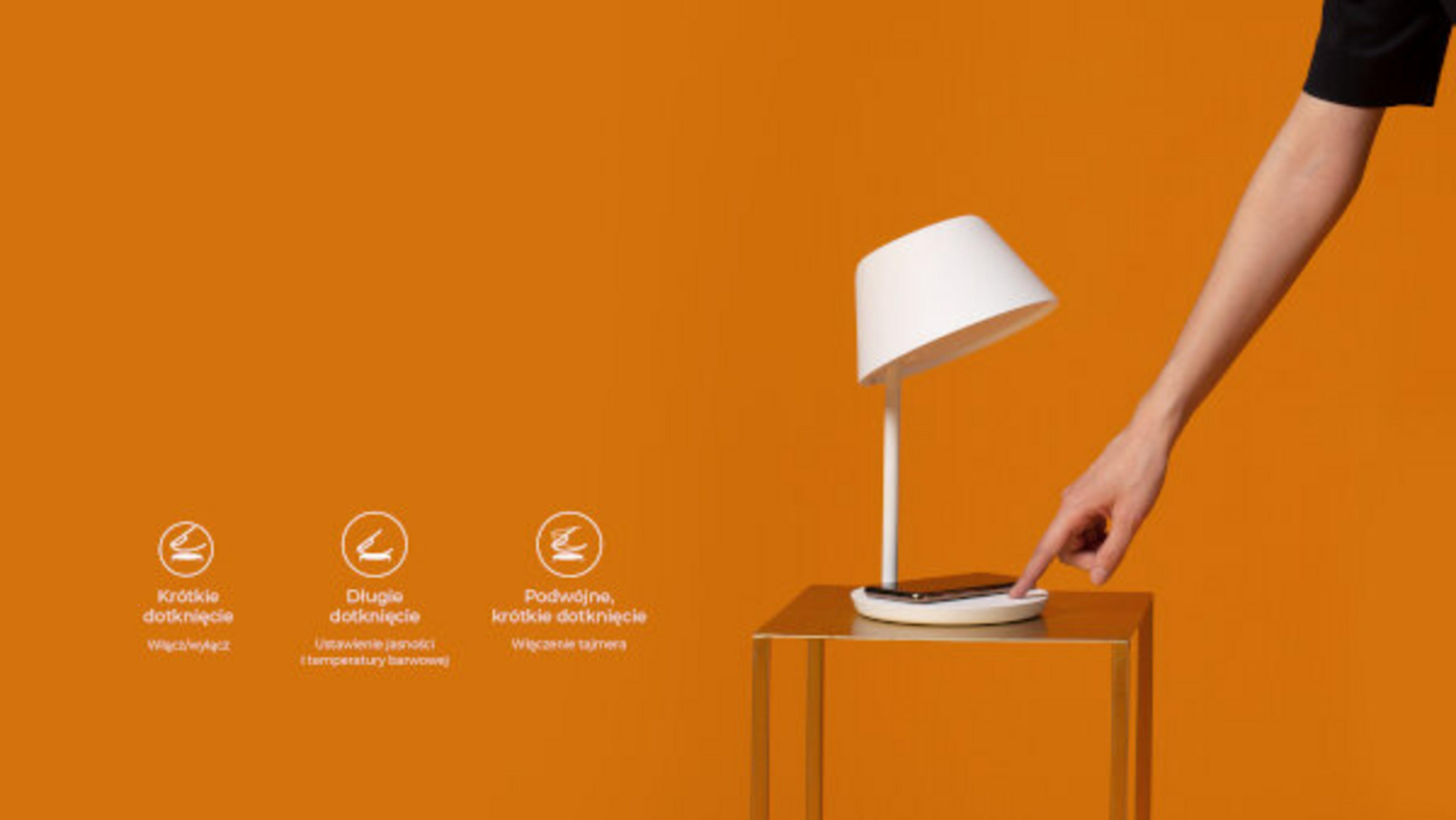 Biała lampa na złotym stoliku. Dłoń dotyka podkładki ładującej. Pomarańczowe tło. Tekst poniżej pokazuje elementy sterujące.