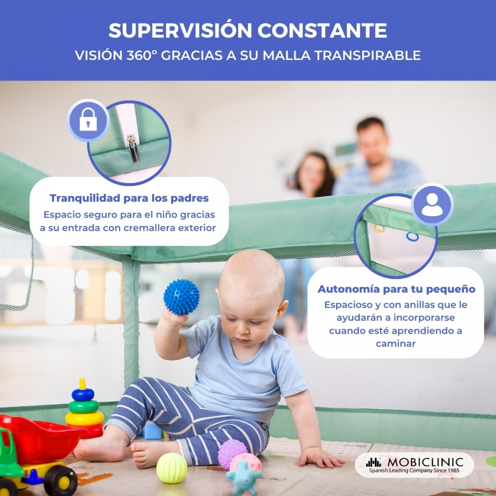 Un bebé juega con juguetes dentro de una cuna. Los padres están borrosos en el fondo. La cuna tiene cremallera y malla.