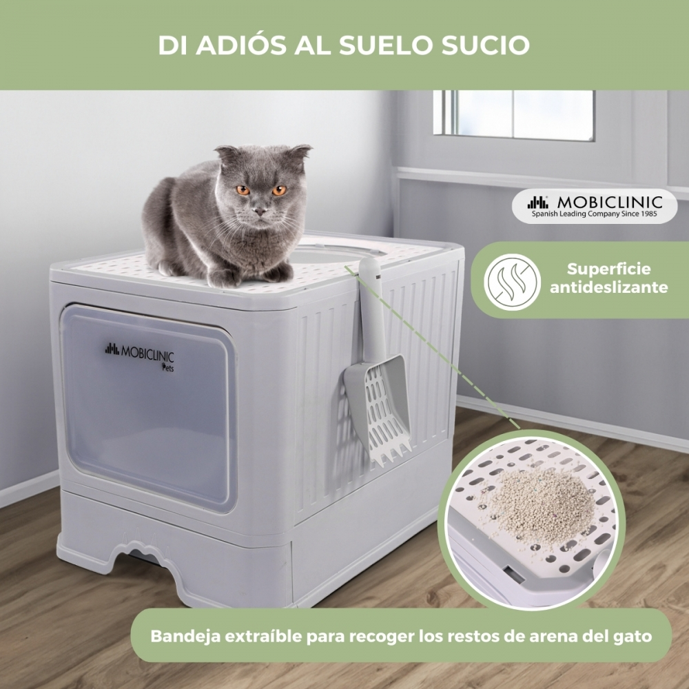 Un gato gris está sentado en una caja de arena blanca. La caja tiene una pala y una bandeja extraíble. El texto en español promociona el producto.