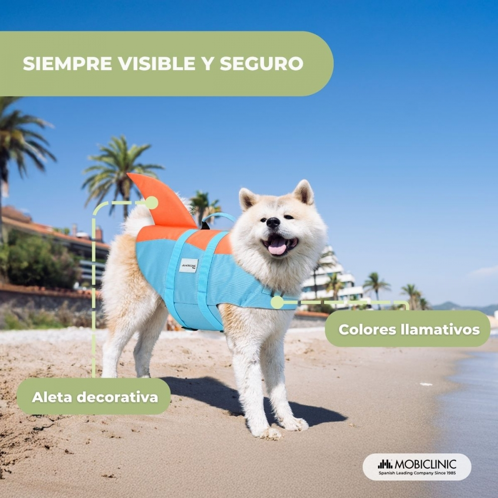 Un perro en la playa lleva un chaleco salvavidas con aleta de tiburón. Está de pie en la arena con el mar y palmeras al fondo.