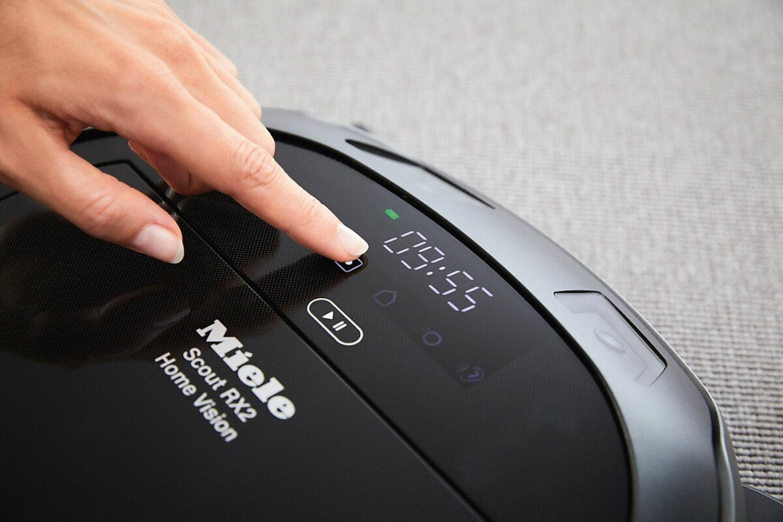Eine Hand drückt eine Taste auf einem schwarzen Miele-Roboterstaubsauger. Das Display zeigt 09:55. Der Hintergrund ist grau.