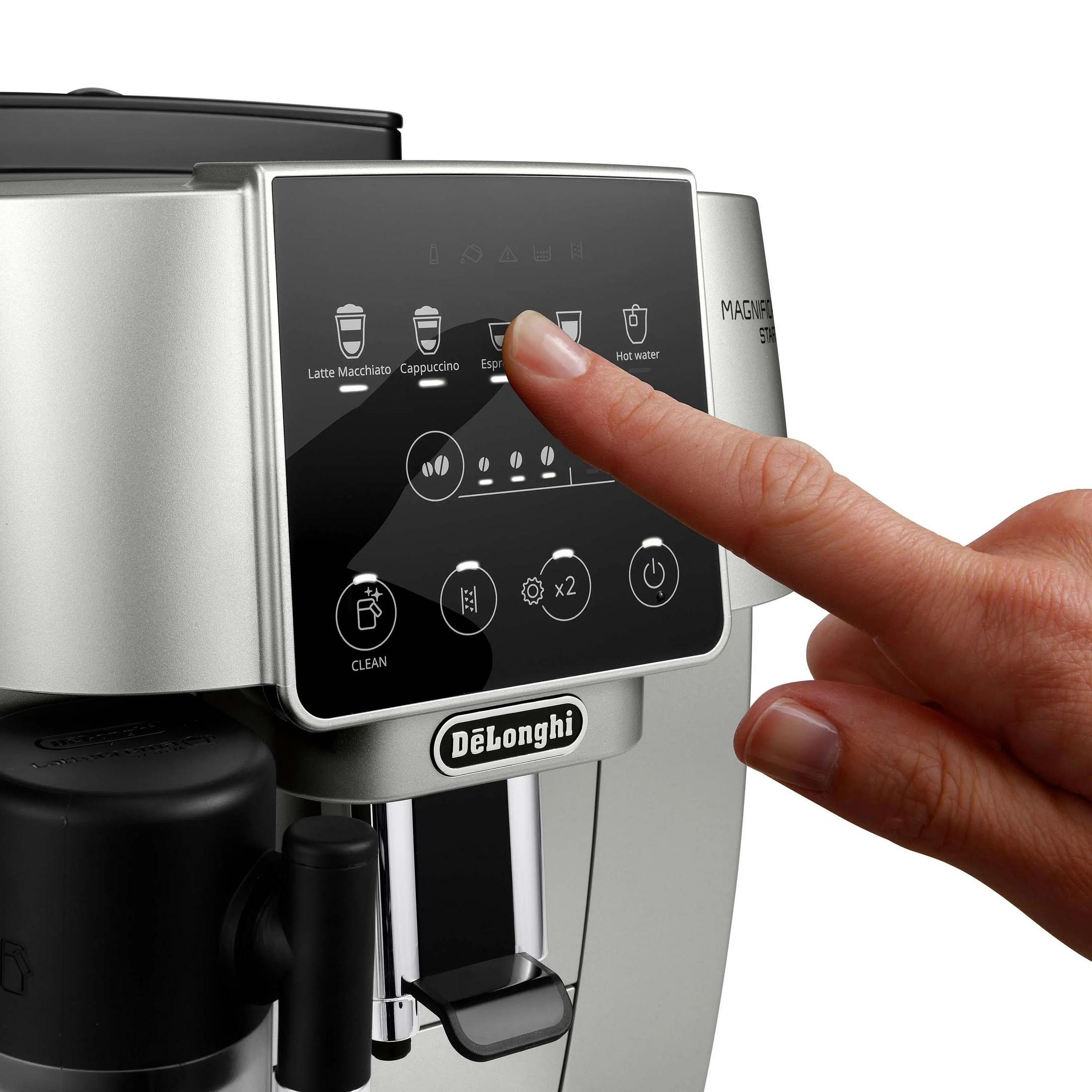 A hand presses a button on a coffee machine. The machine is silver, with a black display showing coffee options.