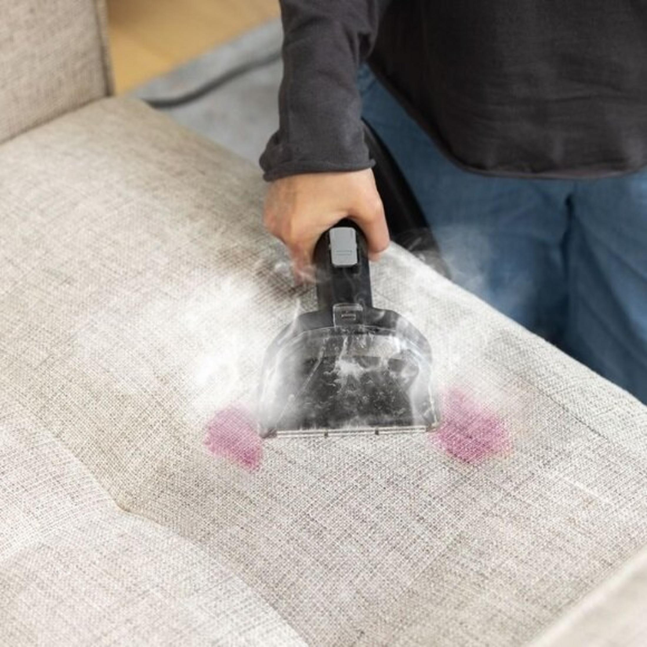 A person cleaning a light-colored sofa with a black upholstery cleaner. Pink stains on the sofa are visible.