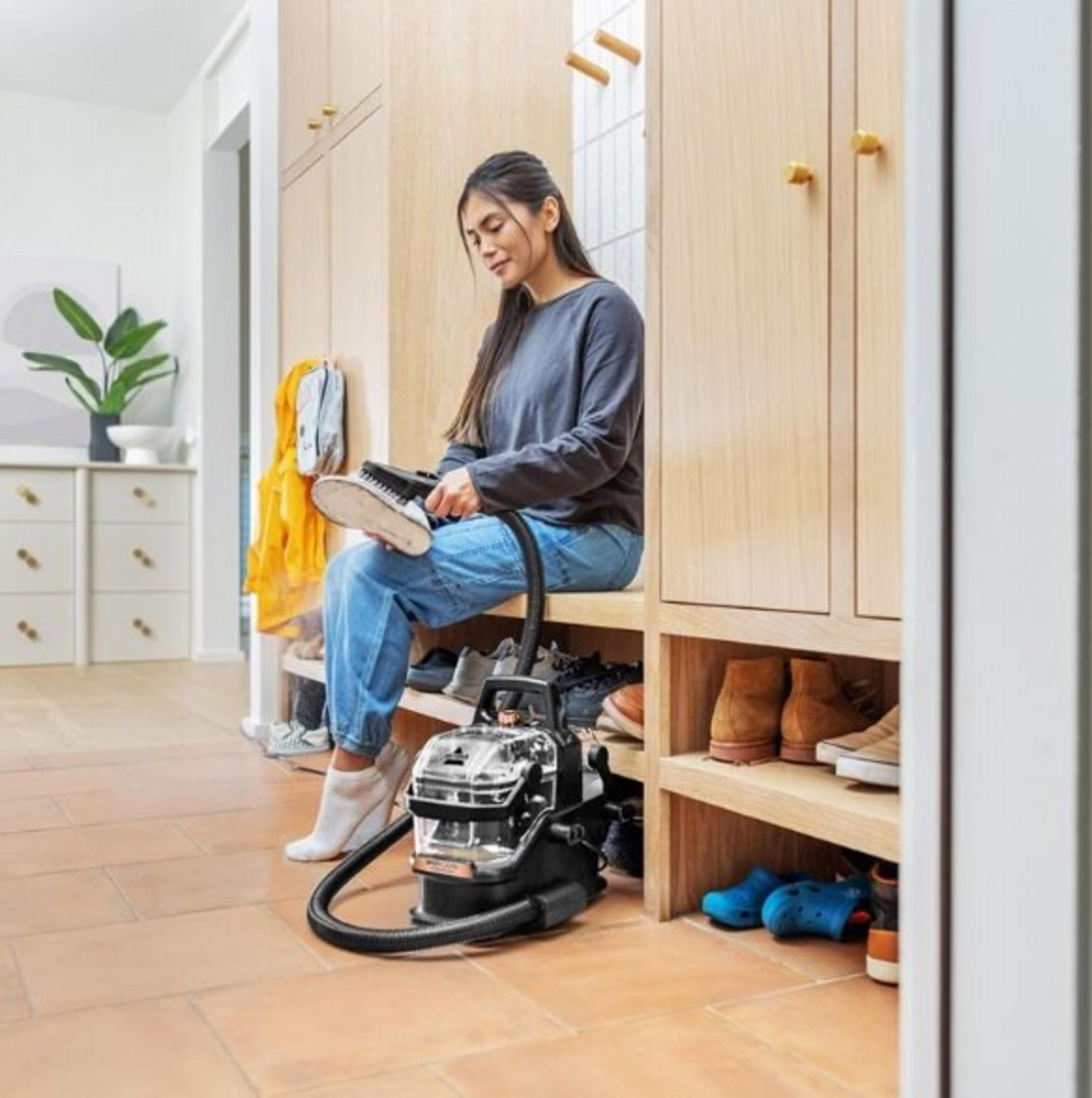 Woman in blue jeans sits, cleaning a shoe. A vacuum cleaner is in front, shoes on shelves, wooden cabinets are in the background.
