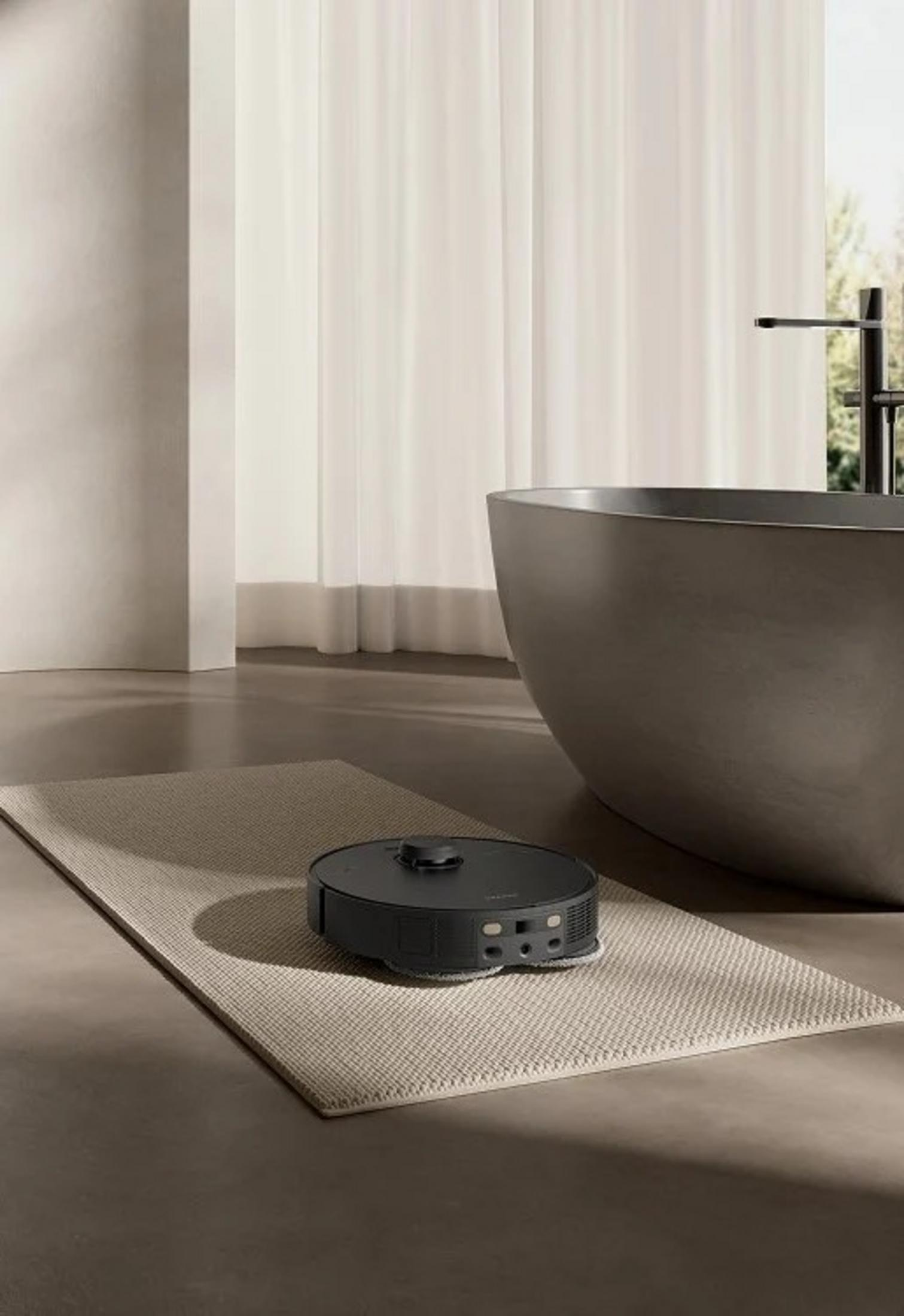 Ciemnoszary odkurzacz automatyczny na beżowym dywanie, przed ciemnoszarą wanną. Białe zasłony i okno w tle.
