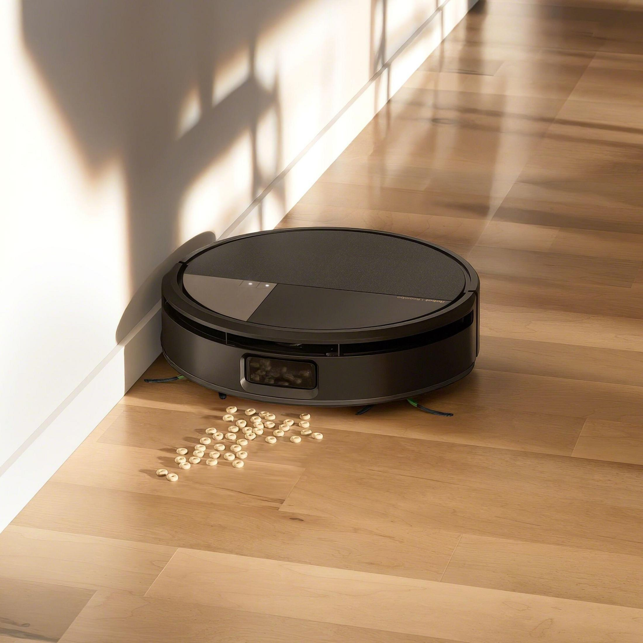 Un robot aspirador gris oscuro sobre suelo de madera, cerca de una pared blanca. Algo de cereal está esparcido.