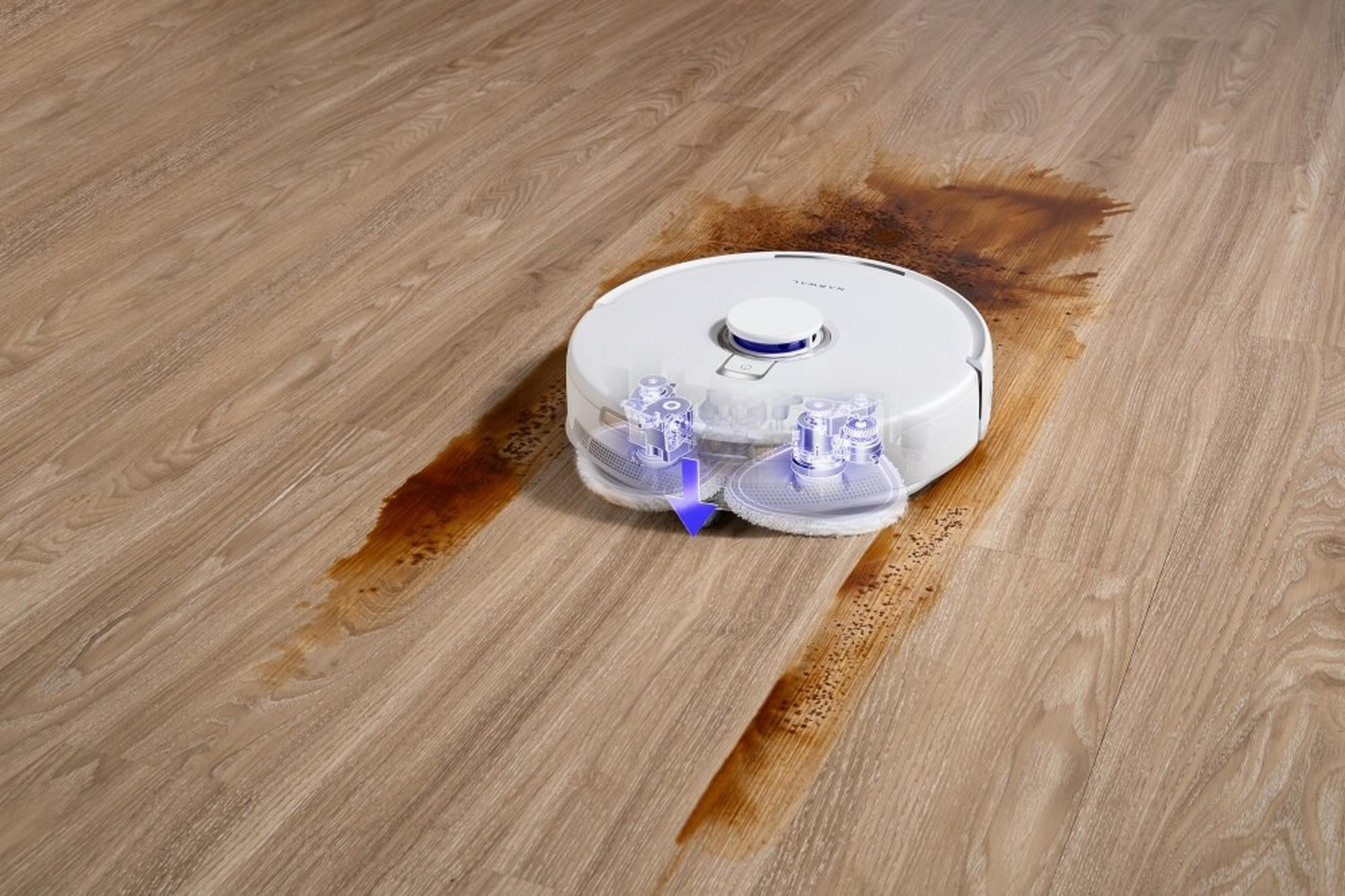 Biały robot sprzątający czyści brązową plamę po płynie na jasnobrązowej drewnianej podłodze.