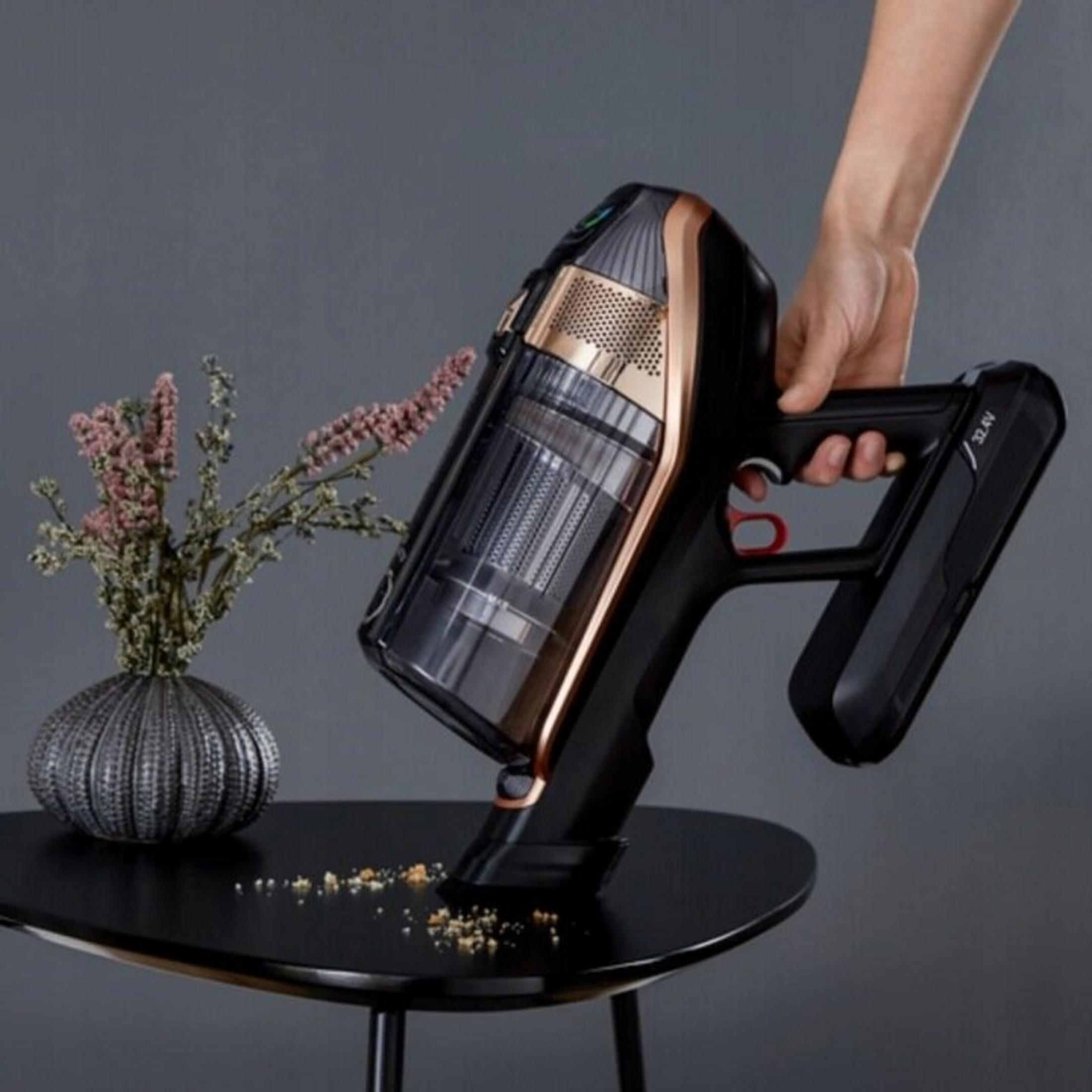 Ręka trzyma czarny i złoty odkurzacz nad czarnym stołem z okruchami. Po lewej stronie wazon z kwiatami.