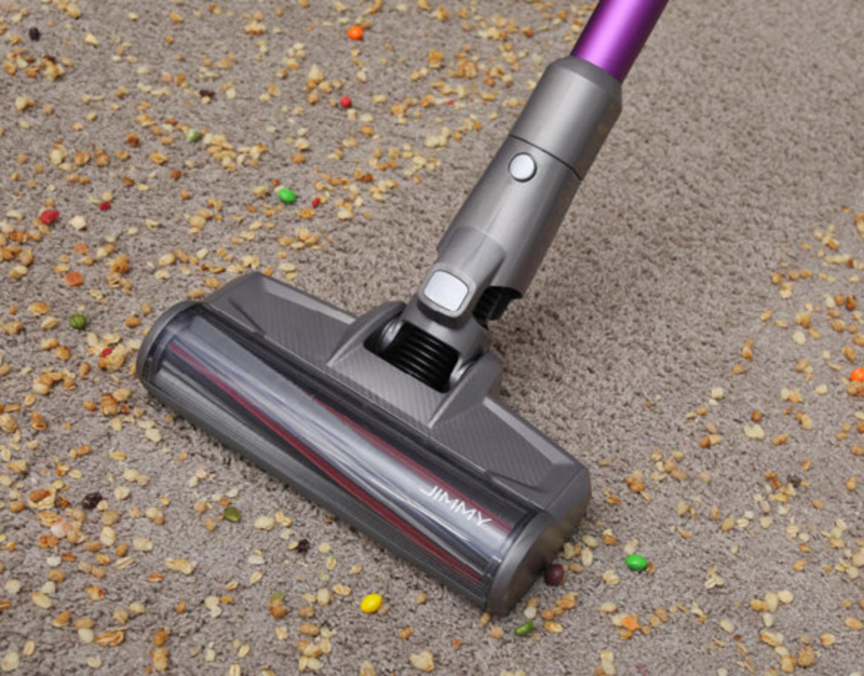 Una aspiradora gris y morada sobre una alfombra con comida y caramelos esparcidos. La aspiradora está en uso.
