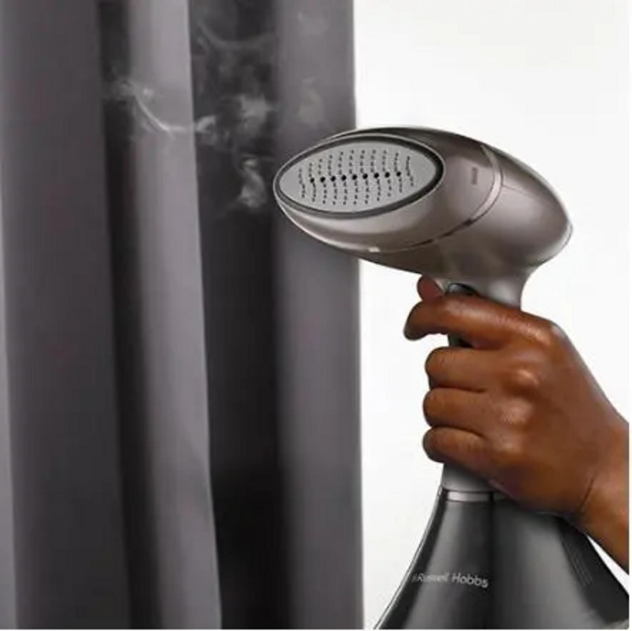 Eine Hand hält einen silbernen Kleiderdampfer in der Nähe grauer Vorhänge. Dampf steigt auf. Der Markenname lautet Russell Hobbs.