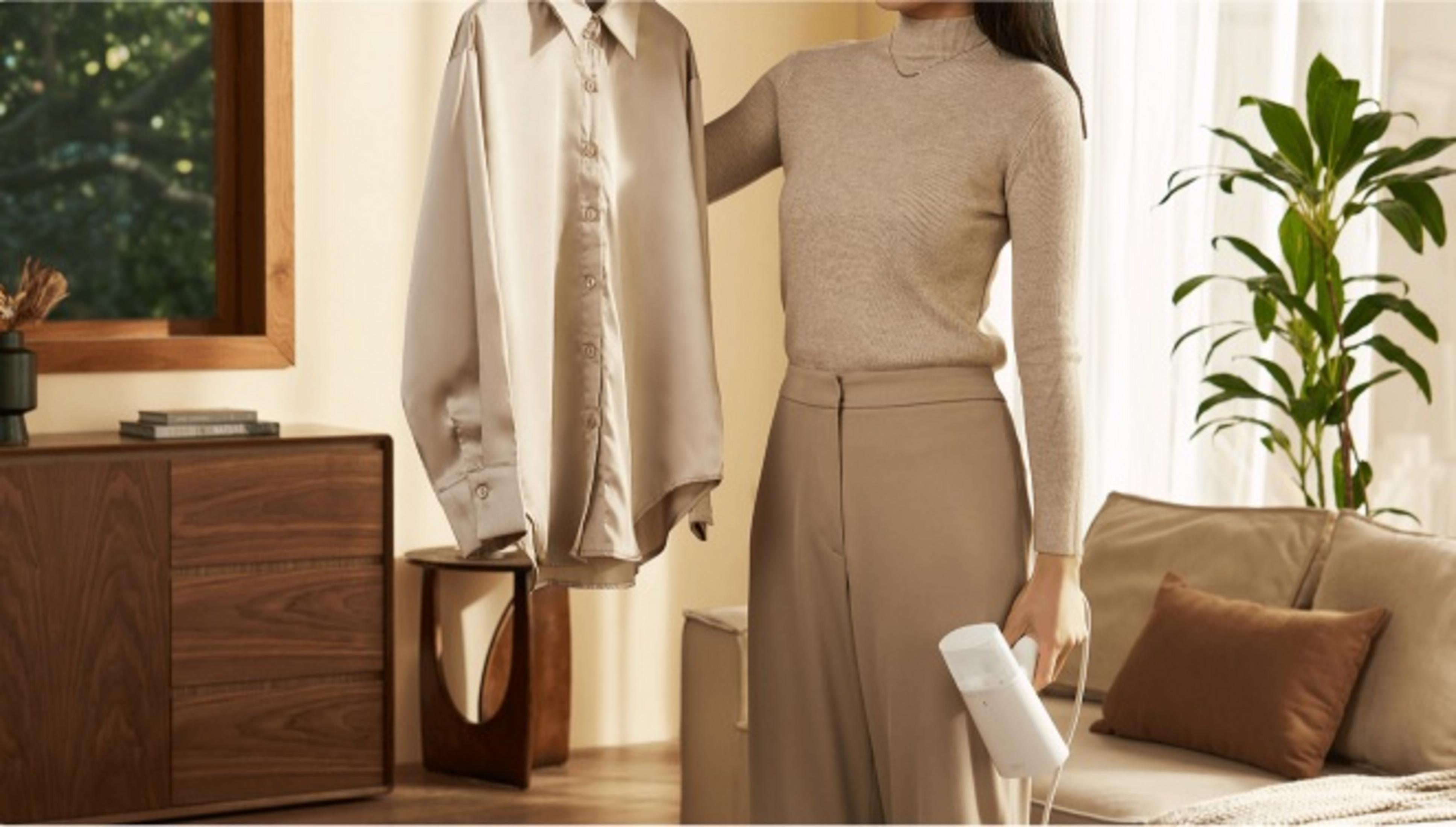 Osoba prasuje koszulę. Trzyma parownicę w jednej ręce, koszulę w drugiej. Roślina i meble są w tle.