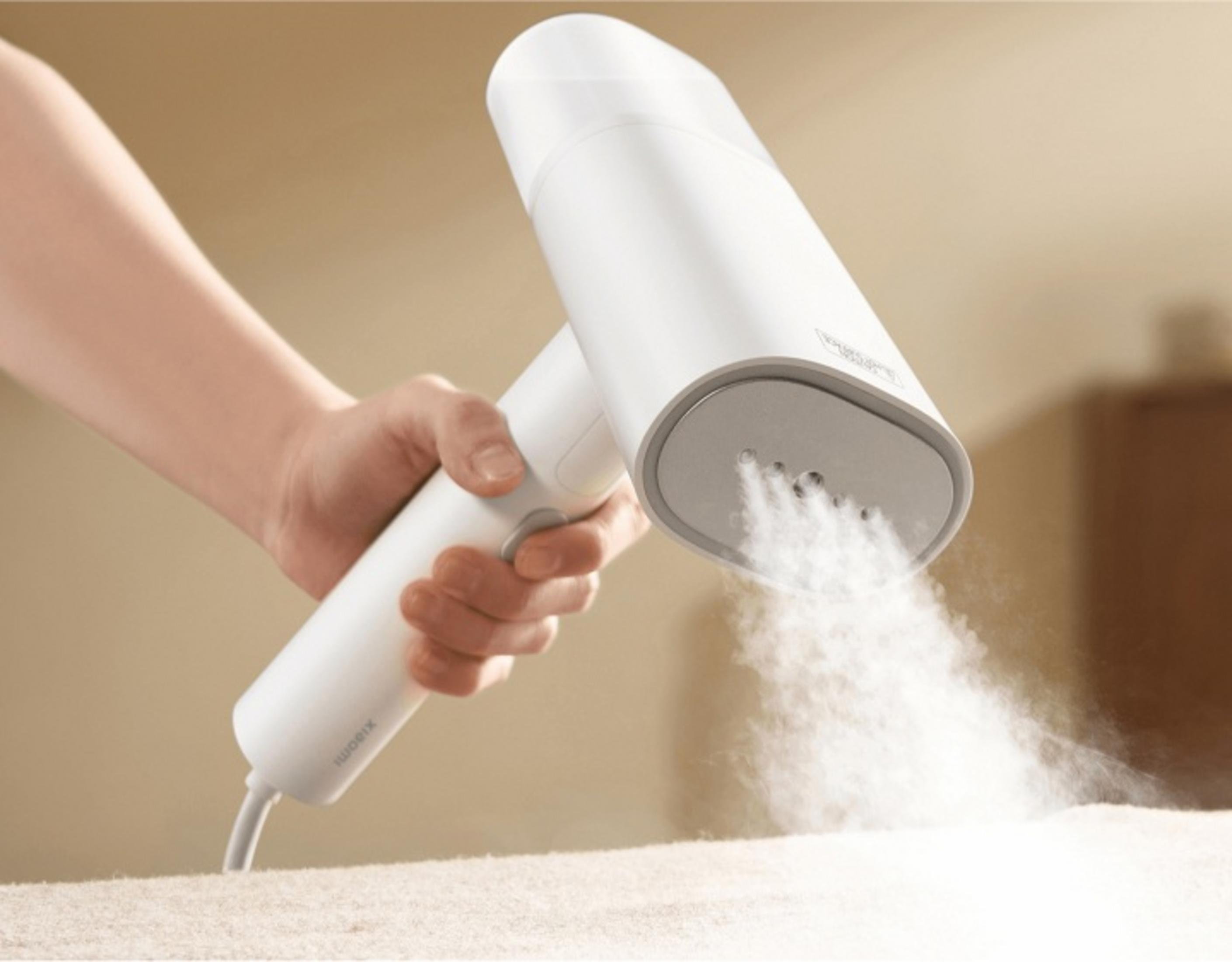 Ręka trzyma białą parownicę do ubrań, uwalniając parę na tkaninę. Beżowe tło.