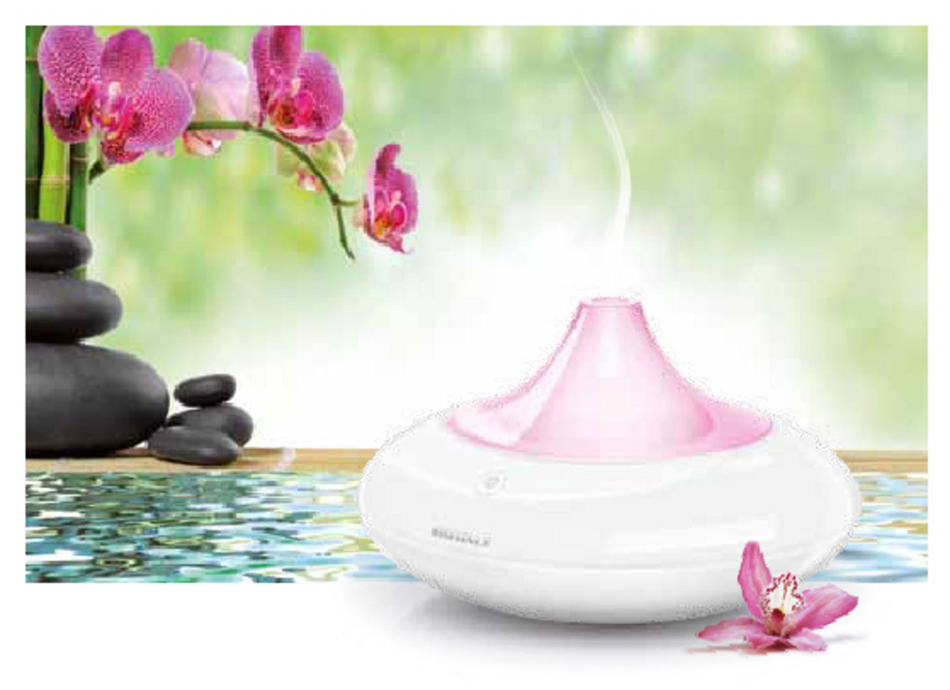 Un diffusore bianco e rosa con orchidee e pietre zen, vapore che sale. Sfondo verde tenue.