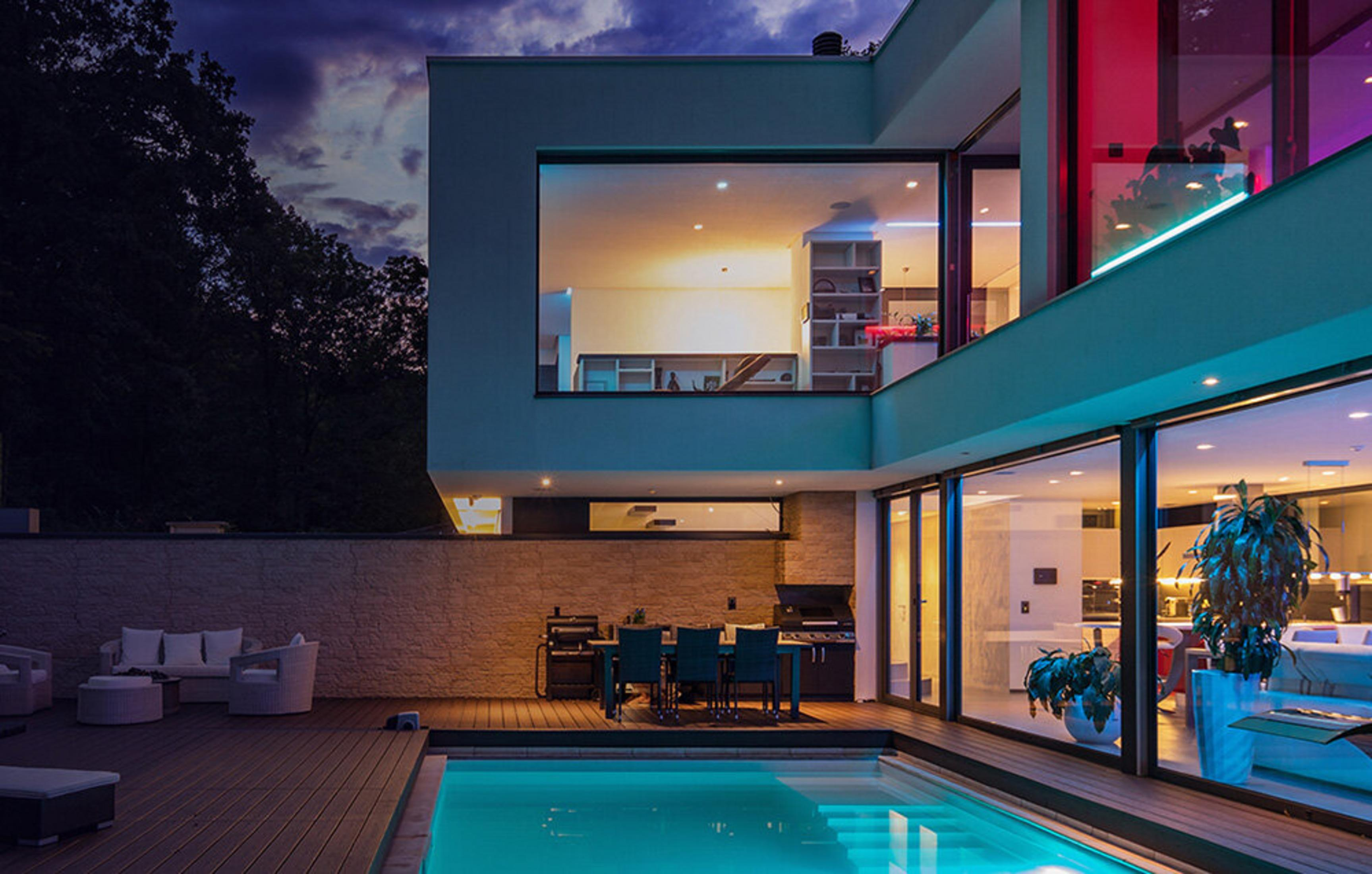 Zewnętrze nowoczesnego domu w nocy. Widoczny basen, patio i duże okna. Drzewa w tle.