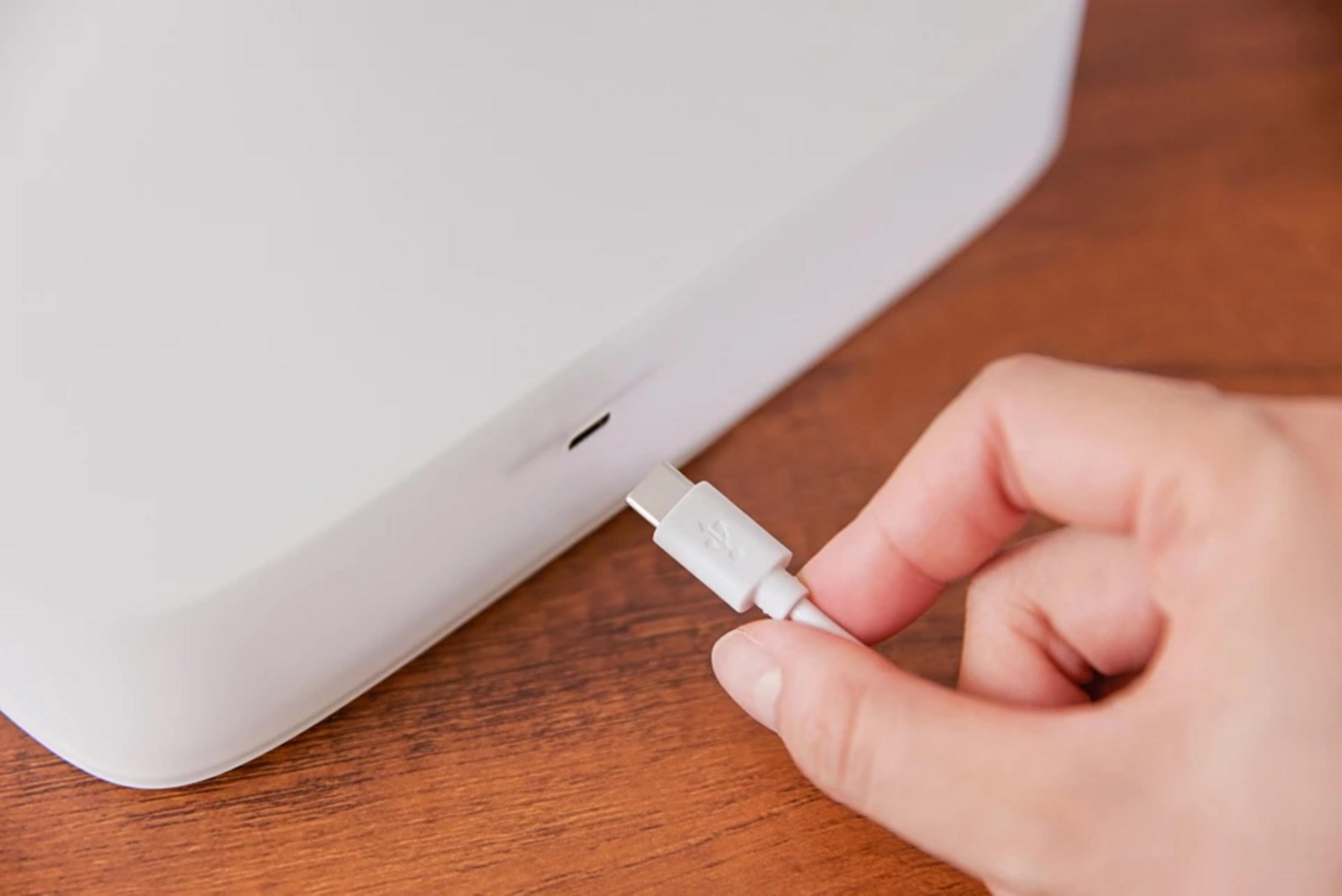 Hand die een witte USB-C-kabel vasthoudt in de buurt van een wit apparaat op een houten oppervlak.