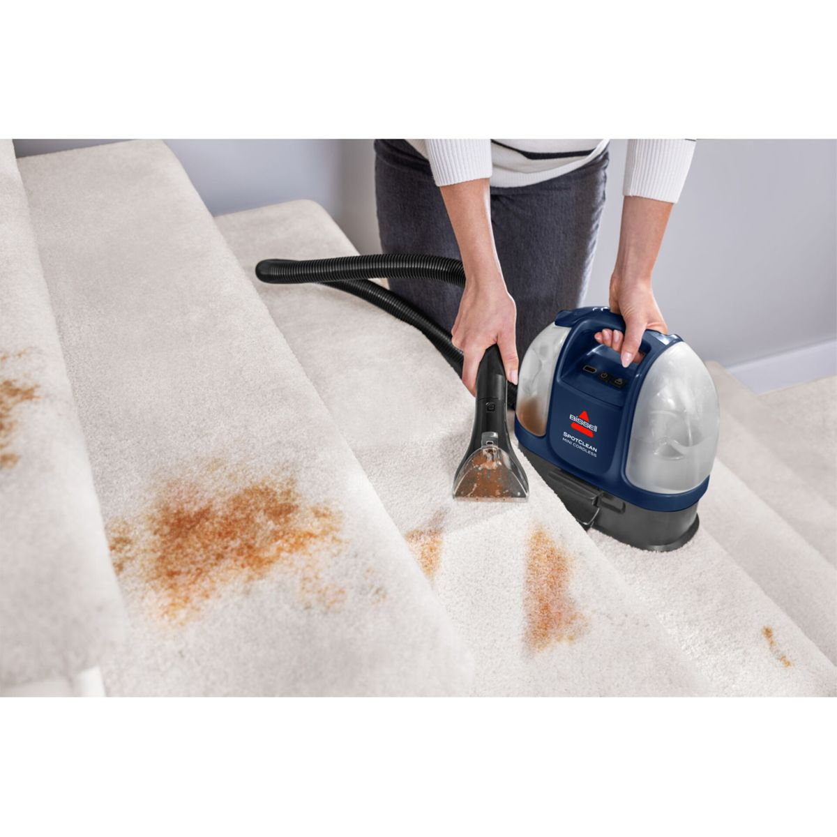 Osoba czyści schody z wykładziną za pomocą odkurzacza Bissell SpotClean. Brązowe plamy są widoczne na kremowym dywanie. Odkurzacz jest niebieski.