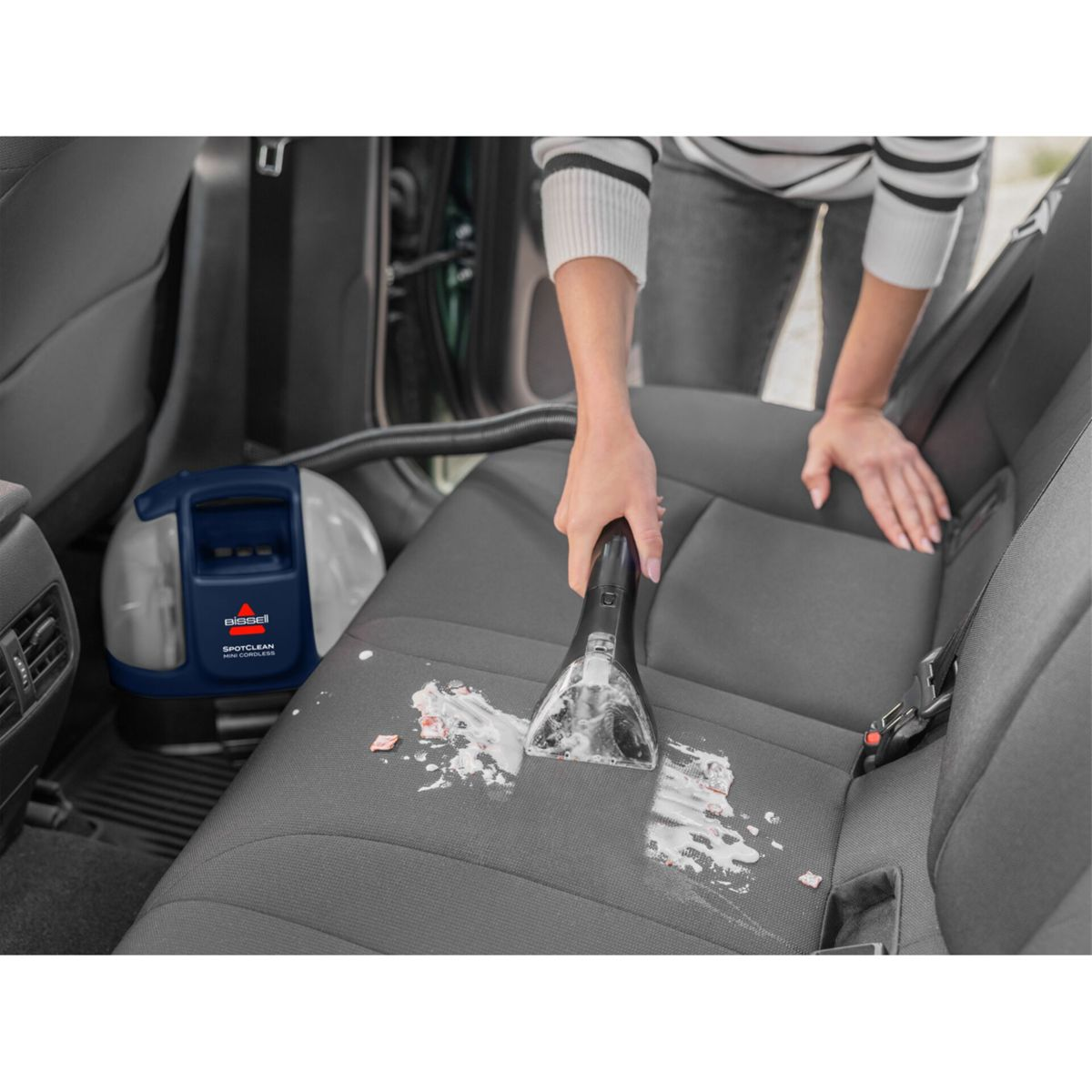 Osoba odkurza siedzenie w samochodzie za pomocą Bissell SpotClean. Siedzenie jest szare i poplamione. W tle znajduje się niebiesko-biały odkurzacz.