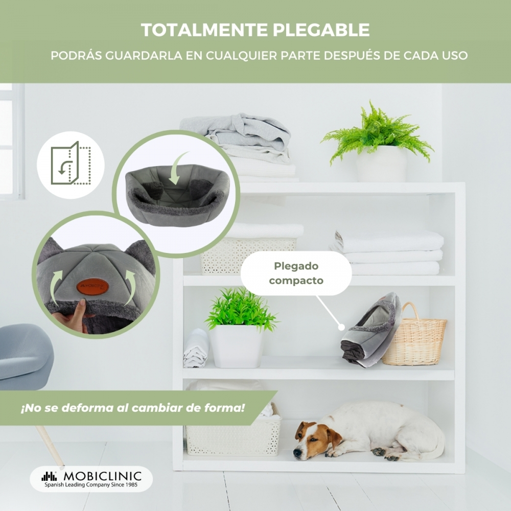 Se muestra una cama plegable para mascotas. Es gris y está doblada. Un perro está acostado cerca de una estantería. La estantería tiene plantas y toallas dobladas.