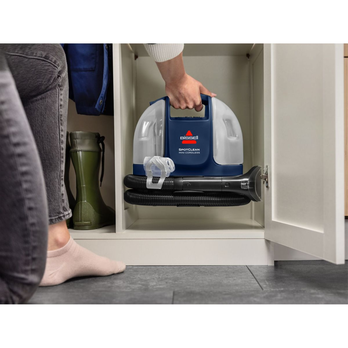 Osoba trzyma Bissell SpotClean Mini Cordless. Znajduje się w szafce z niebieską kurtką i zielonymi butami.