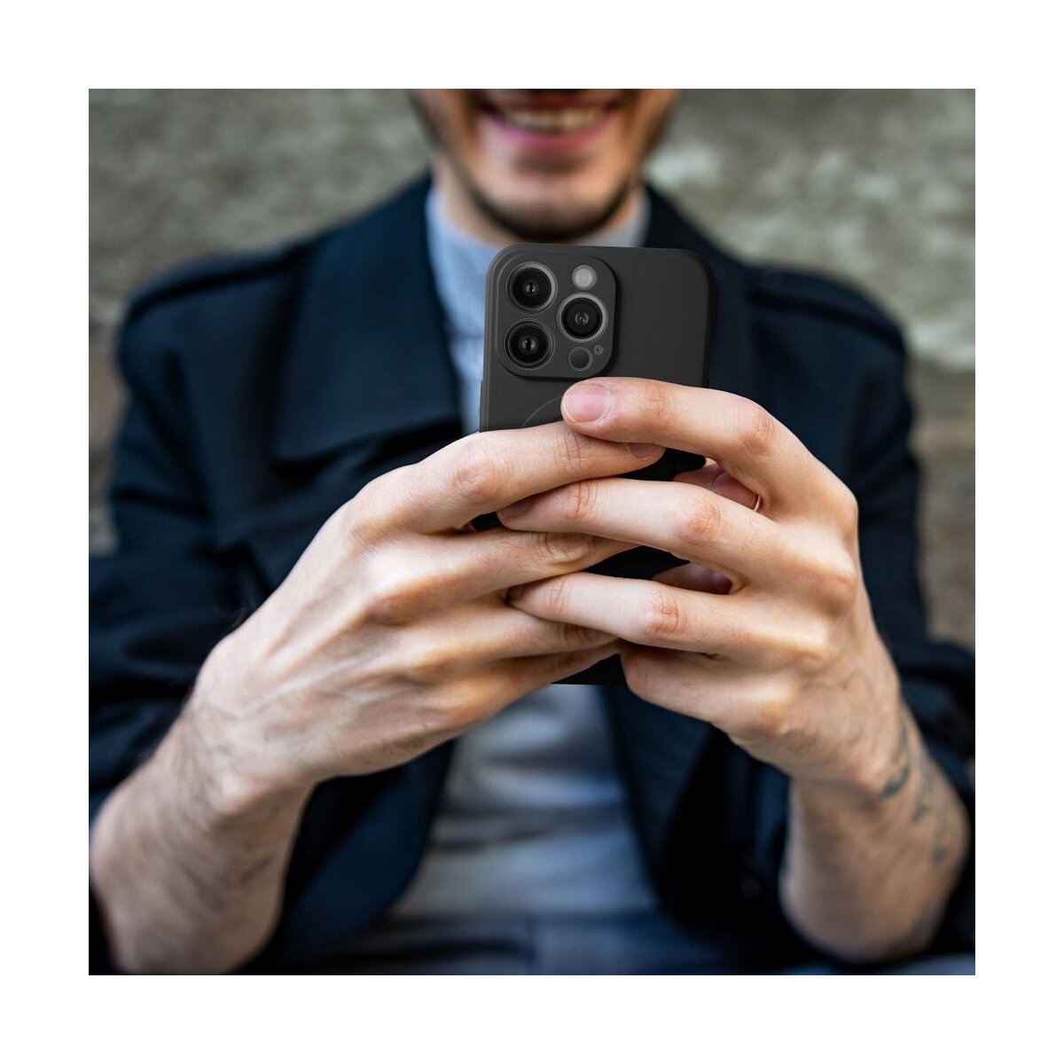Eine Person hält ein schwarzes Telefon. Das Telefon hat drei Kameras. Die Person lächelt. Der Hintergrund ist eine verschwommene Wand.