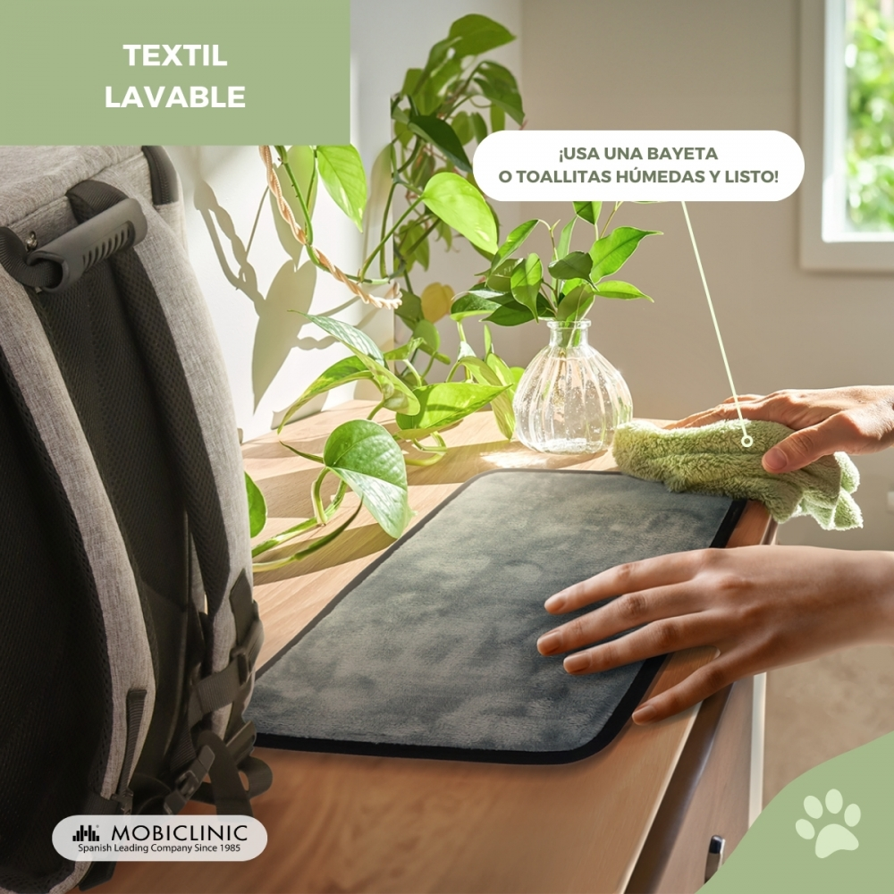 Una alfombra gris se limpia con un paño verde sobre una mesa de madera. Una mochila está a la izquierda, plantas al fondo.