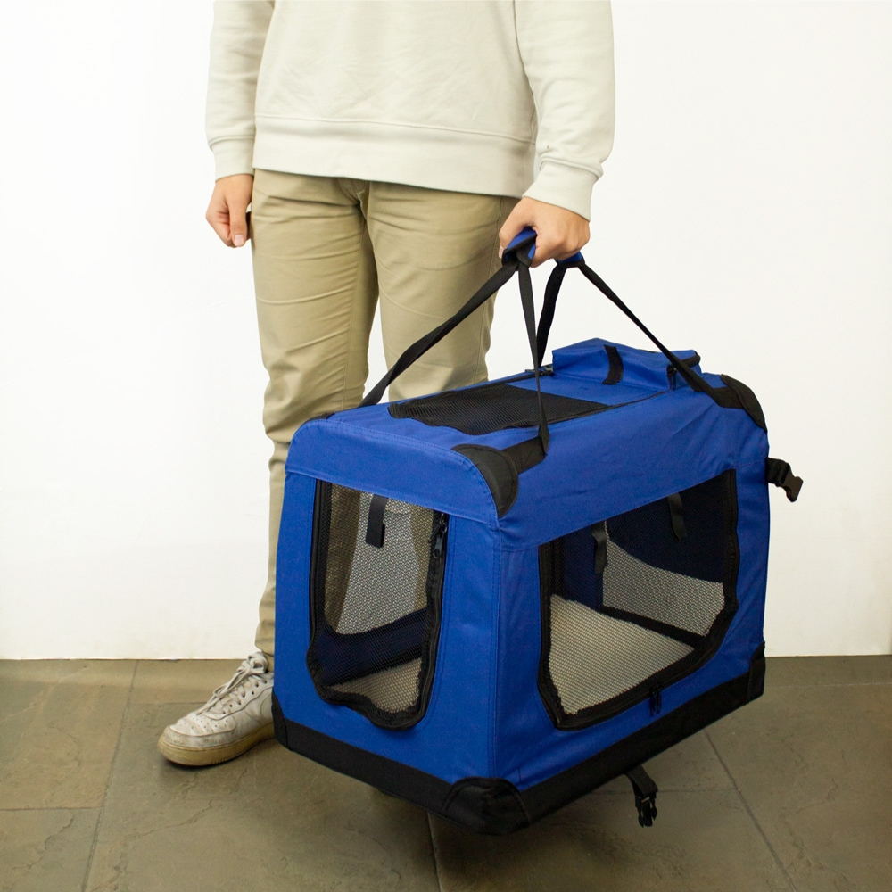 Una persona sostiene un transportín azul para mascotas. El transportín tiene ventanas de malla y correas negras. La persona lleva pantalones beige y una camisa blanca.