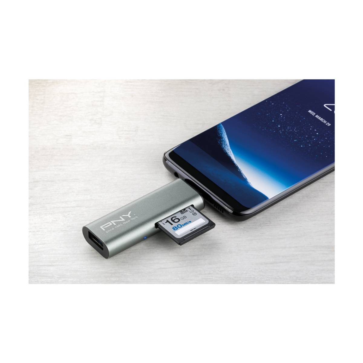 Un teléfono conectado a un lector de tarjetas con una tarjeta SD insertada. La pantalla del teléfono muestra una galaxia azul. El lector es plateado.