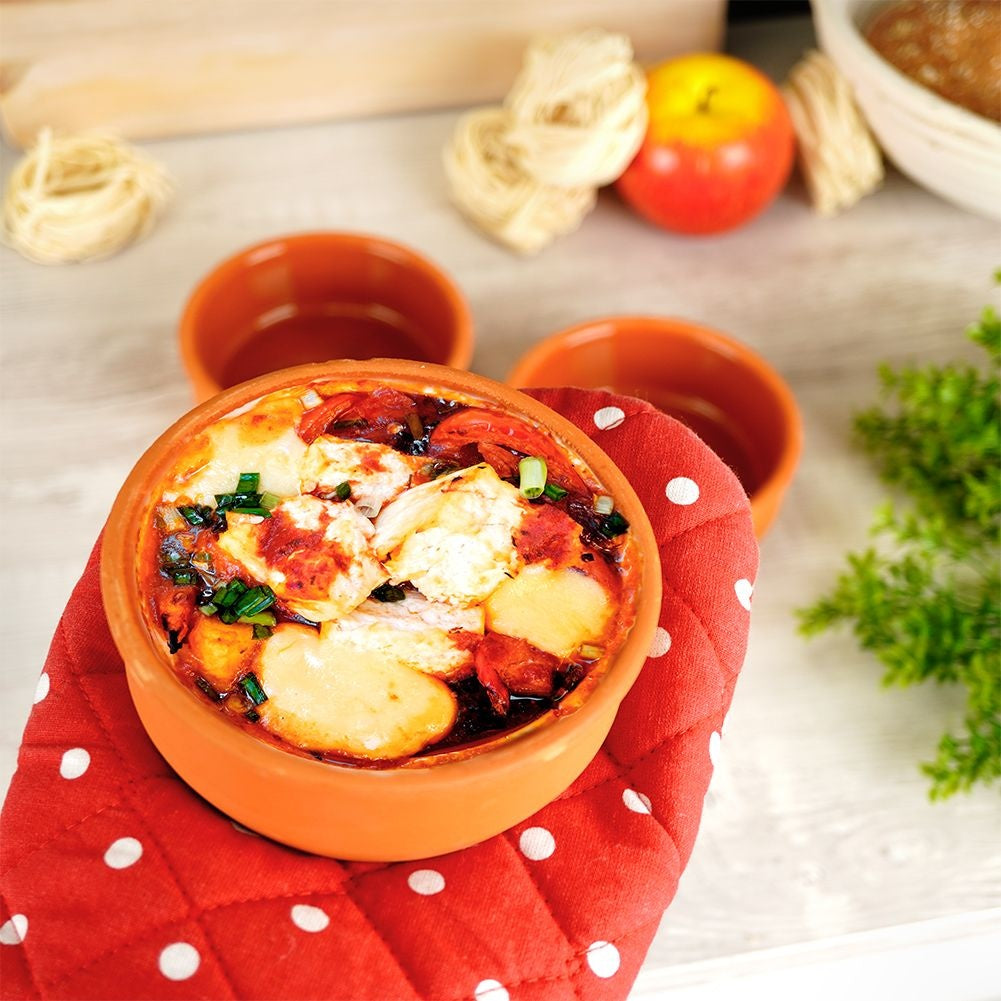 Piatto al forno in ciotola arancione su guanto rosso a pois. Contiene formaggio fuso, pomodori e cipolline. Altre ciotole e cibo sullo sfondo.