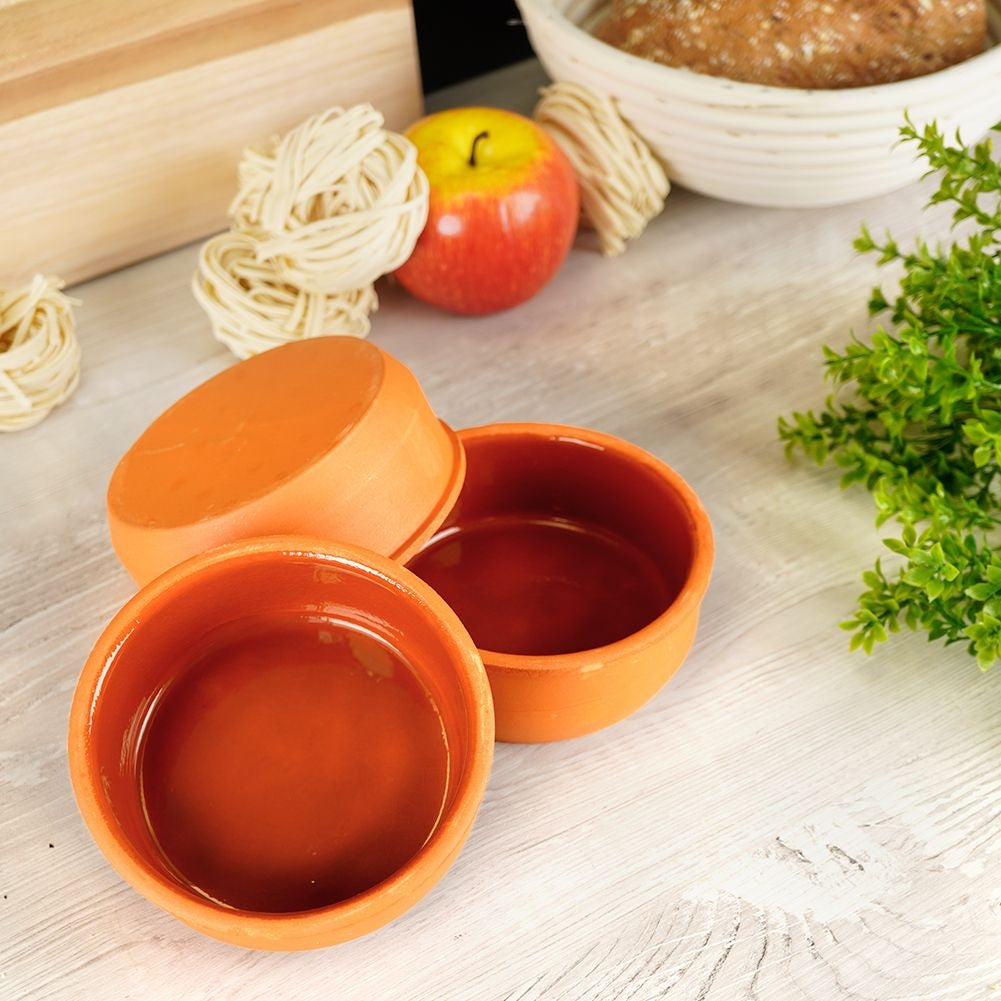 Tre ciotole in ceramica arancione, una mela rossa, nidi di pasta e pane in un cestino su una superficie di legno bianca.