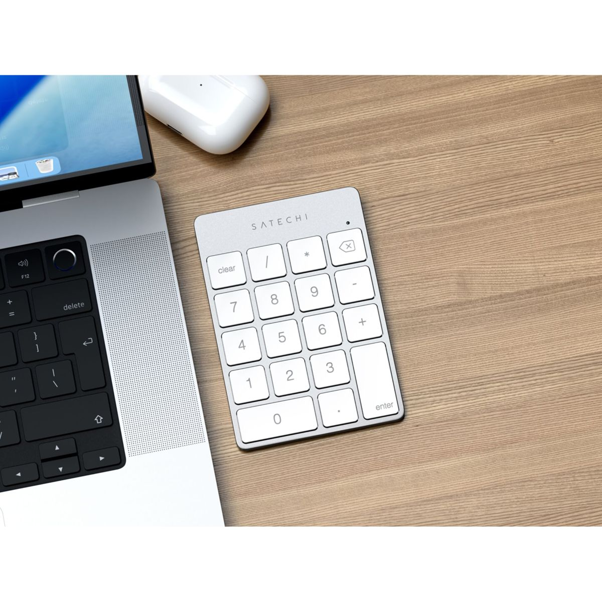 Un teclado numérico plateado con teclas blancas está sobre un escritorio de madera junto a una laptop y auriculares inalámbricos. El teclado numérico está etiquetado Satechi.