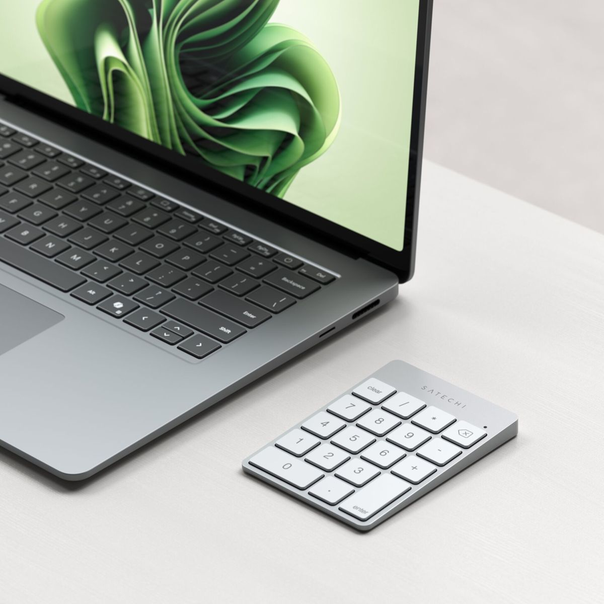 Laptop y teclado numérico en superficie blanca. El laptop muestra arte abstracto verde. El teclado numérico tiene teclas blancas y marco plateado.