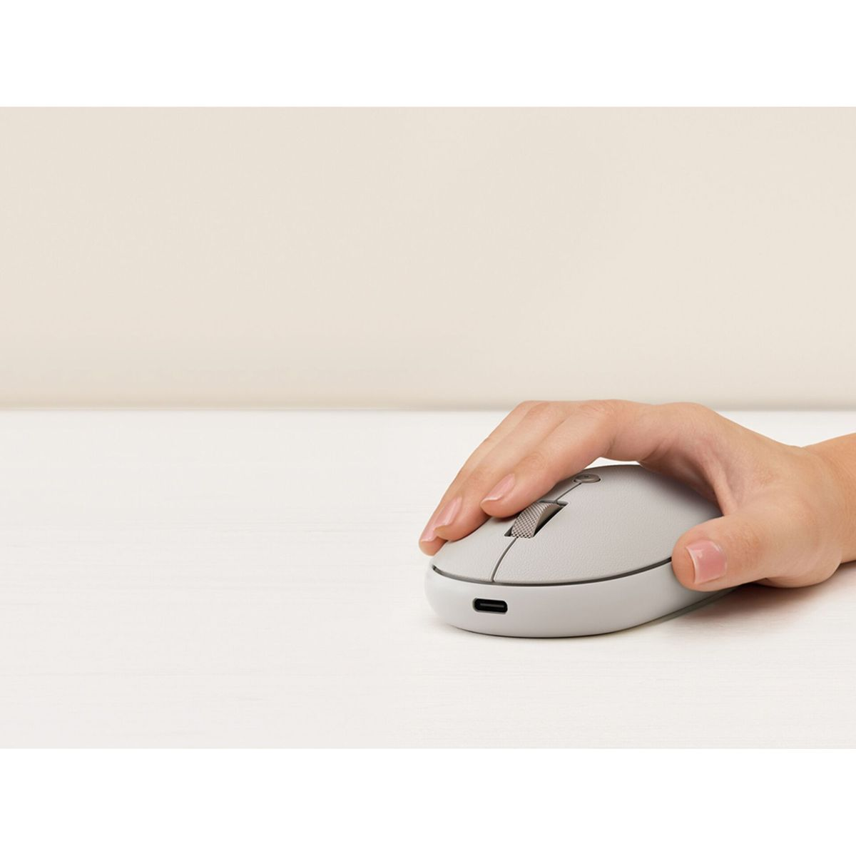 Una mano è appoggiata su un mouse grigio. Il mouse è su una superficie bianca. Lo sfondo è un muro beige chiaro.